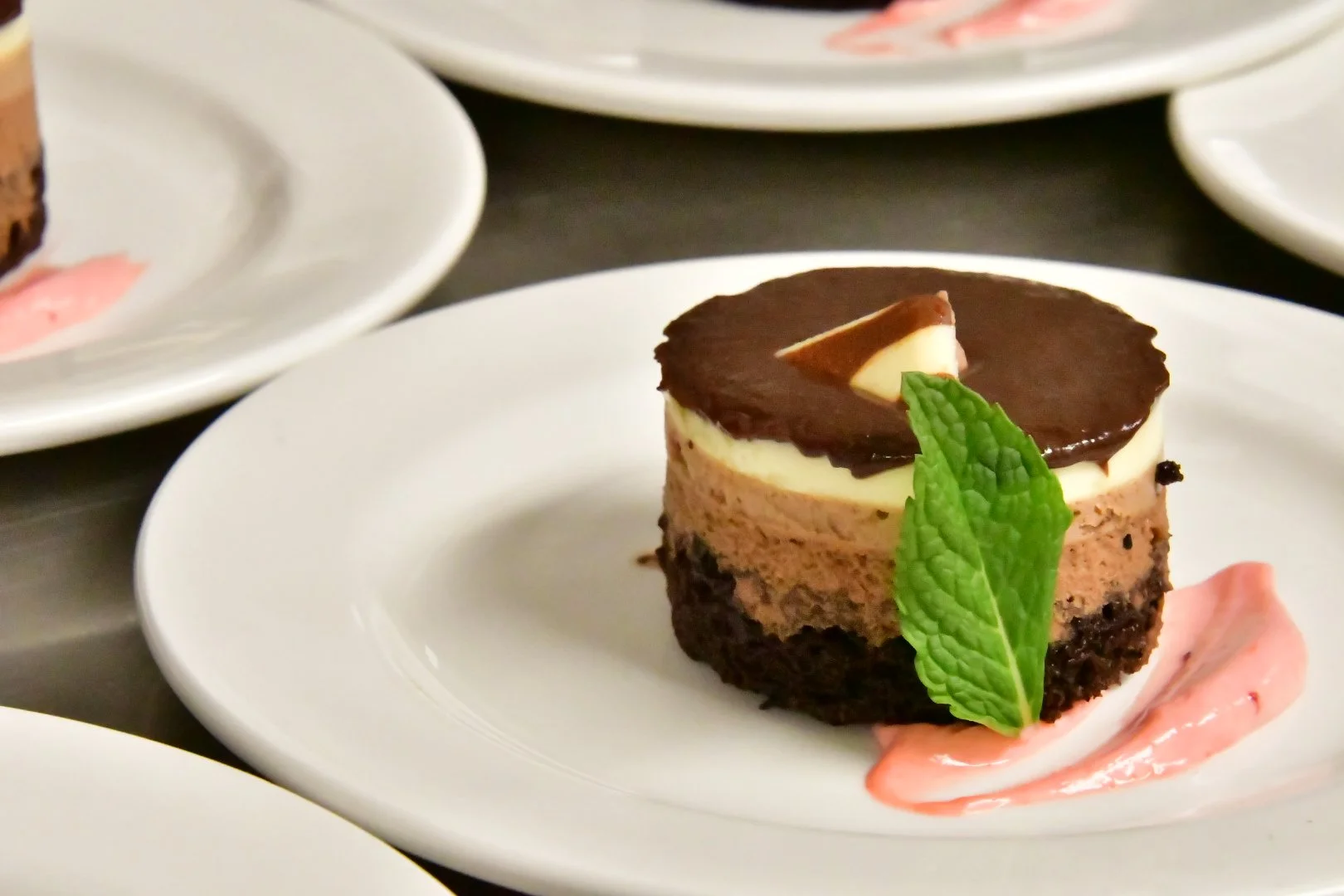 Layered chocolate mousse dessert garnished with a mint leaf on a white plate, with a pink cream decoration on the side.
