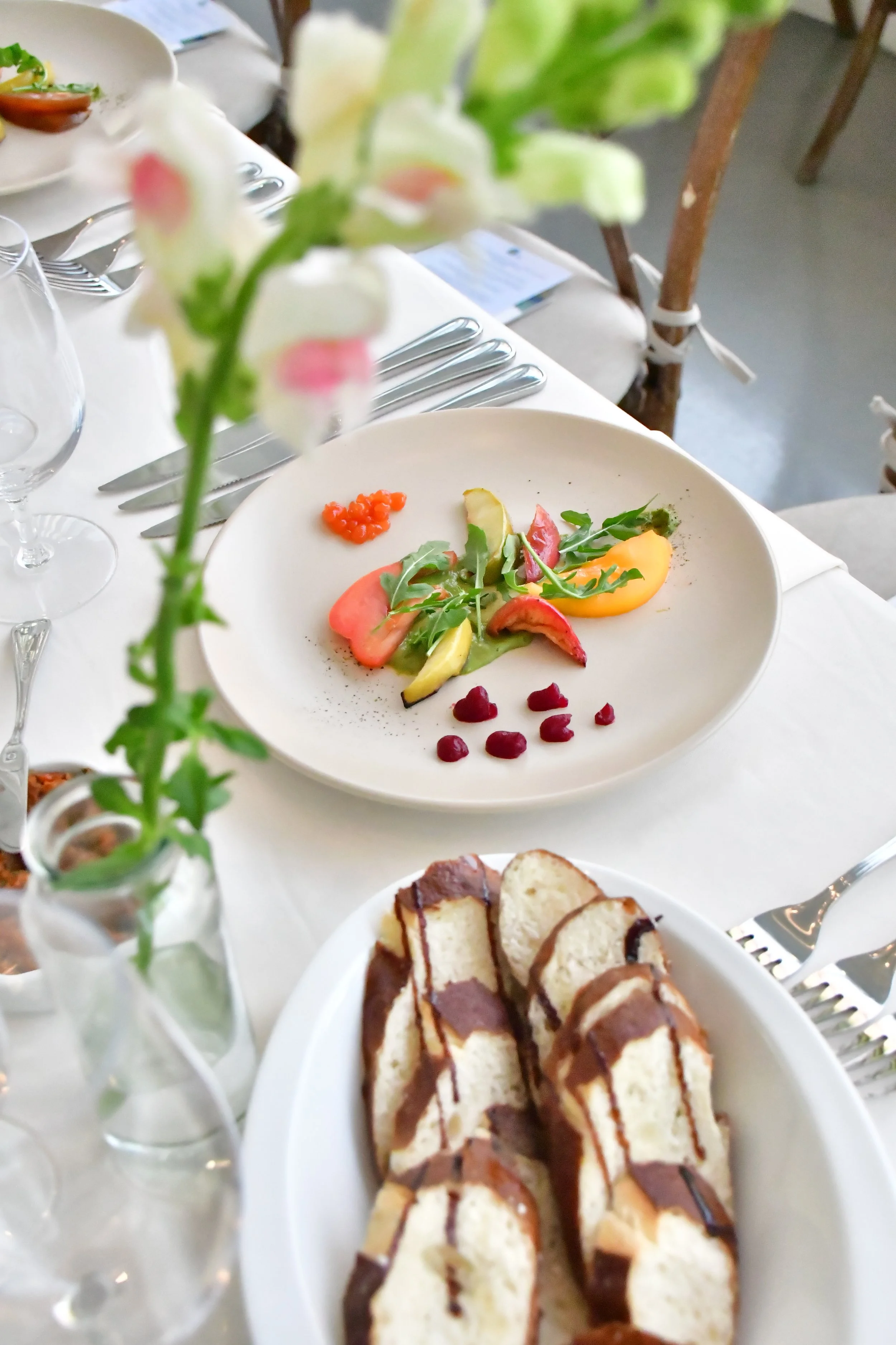 Elegant table setting with flowers in bud vases set with plates of colourful salads, including a bowl of sliced soft pretzel baguette drizzled with balsamic glaze, on a white tablecloth with glassware and silverware.