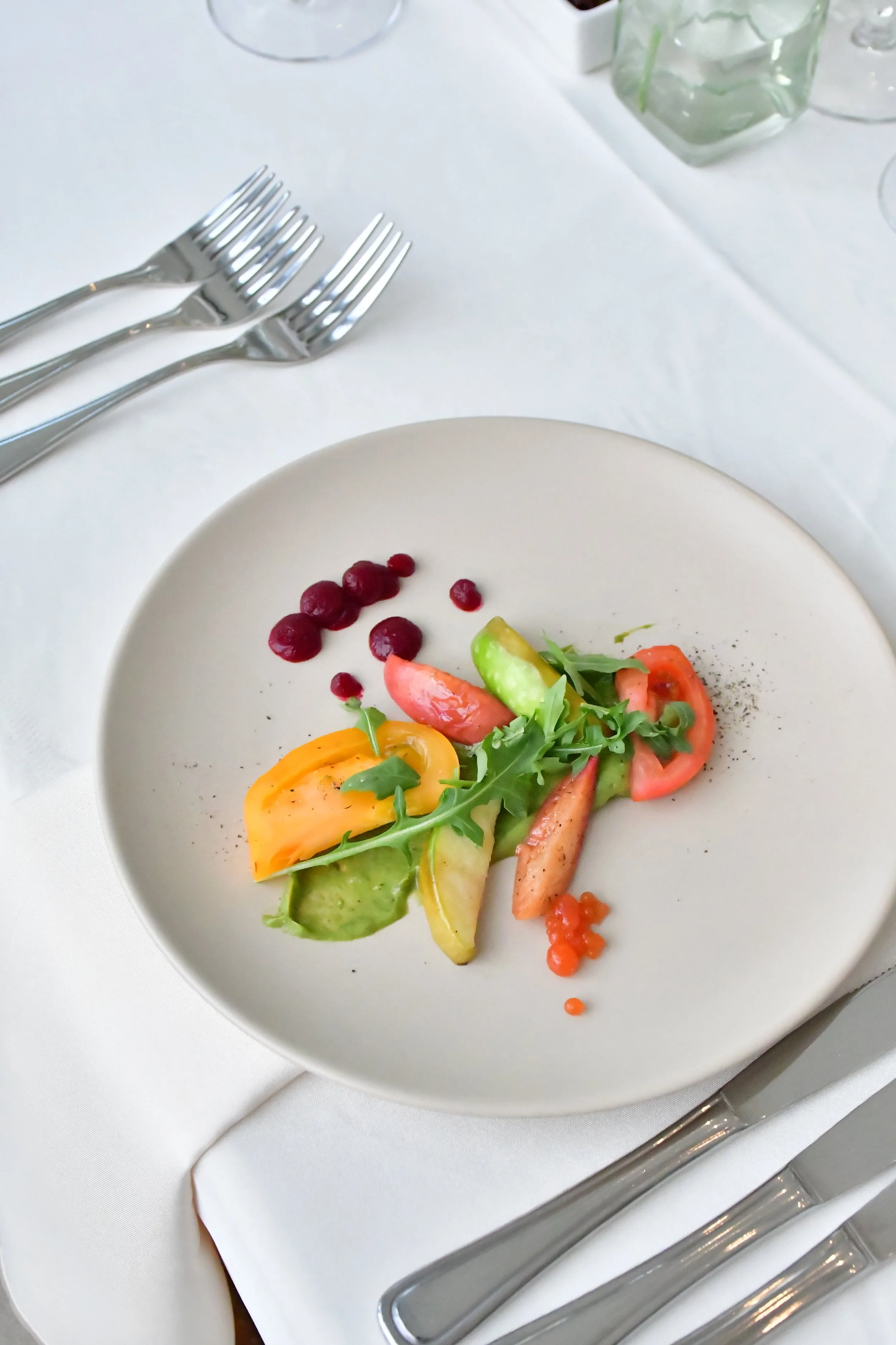 A white plate with a small, colourful salad consisting of heirloom tomatoes, arugula, marinated pears, garnished with black pepper, and tomato caviar pearls on a table-scape set with formal silverware setting, and white tablecloth.