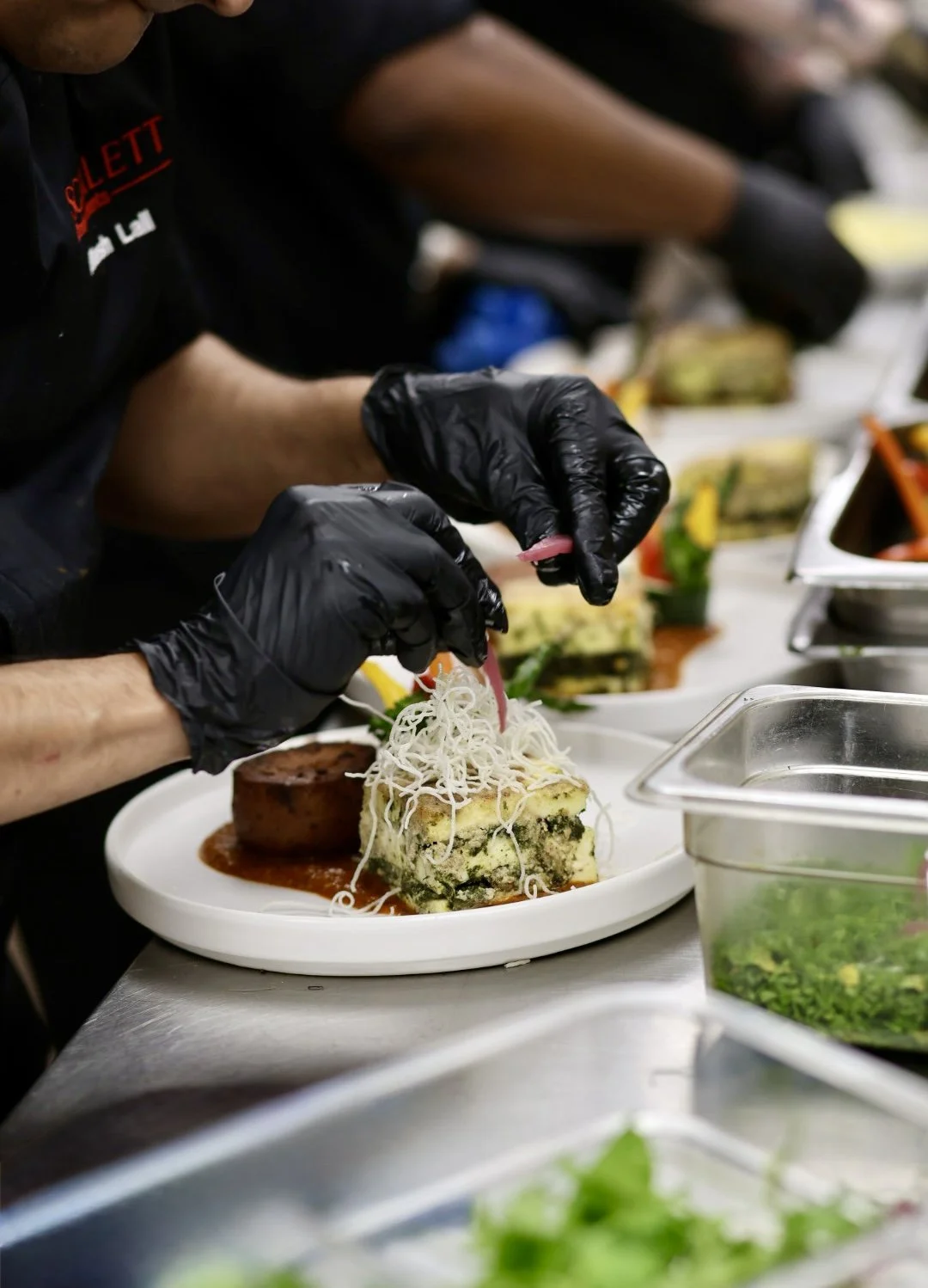 A chef wearing black gloves and a black shirt garnishes a plated dish with shredded cheese and a pink ingredient at a busy restaurant kitchen.