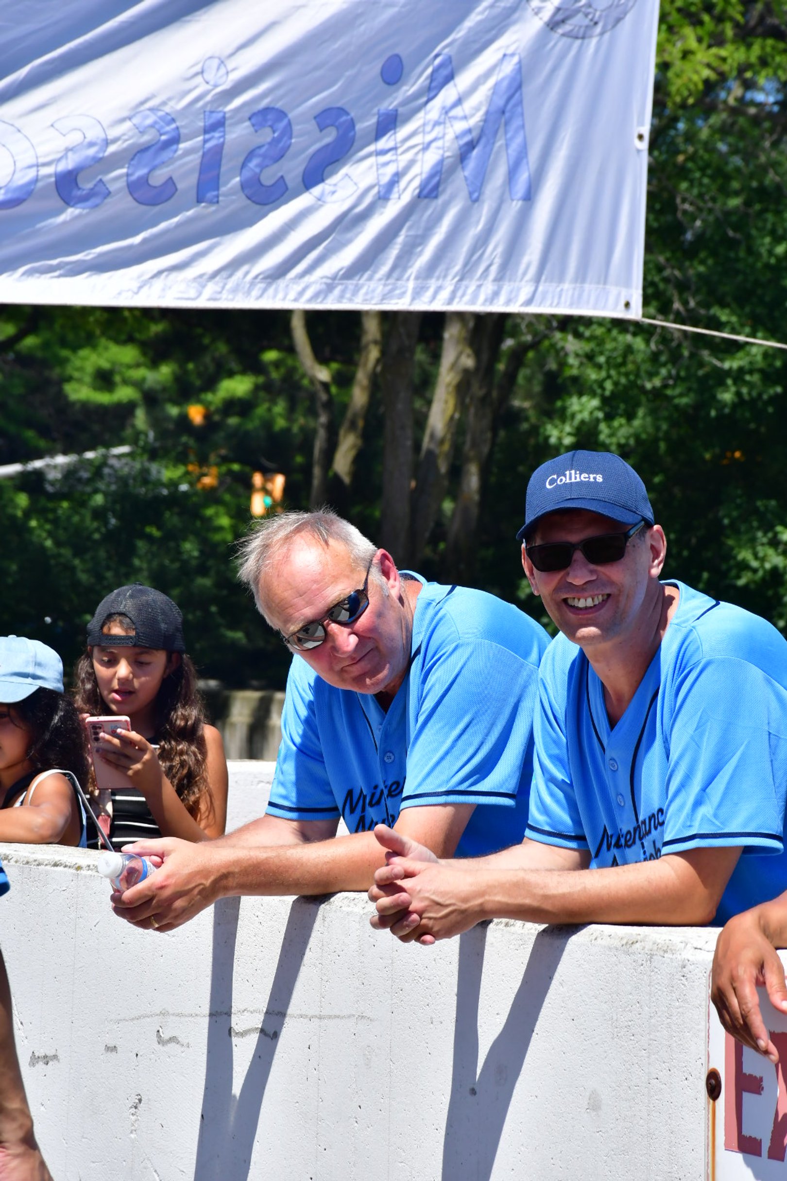 Two men and two young girls at a public event outdoors, with trees in the background. The men are wearing matching blue shirts and hats, and the girls are in casual clothing with one wearing a cap and the other a hat.