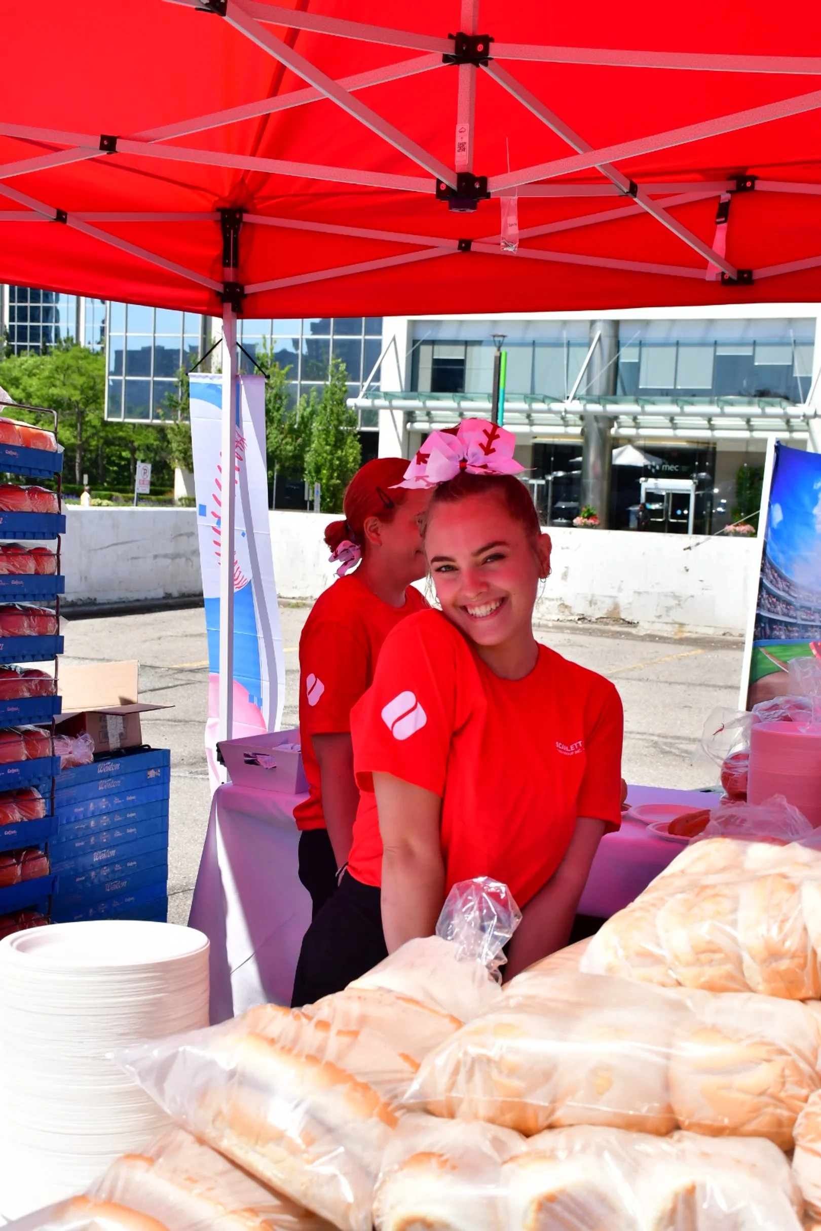 A woman wearing a red scarlett house shirt and baseball-themed headband, standing behind a table with fresh baked buns, under a red canopy at an outdoor event.