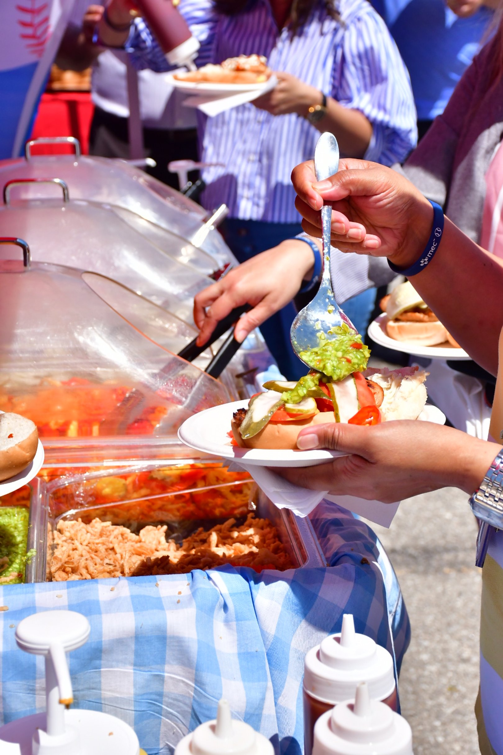 People serving themselves food from a buffet with toppings like relish and tomatoes, during an outdoor event with multiple people in the background.
