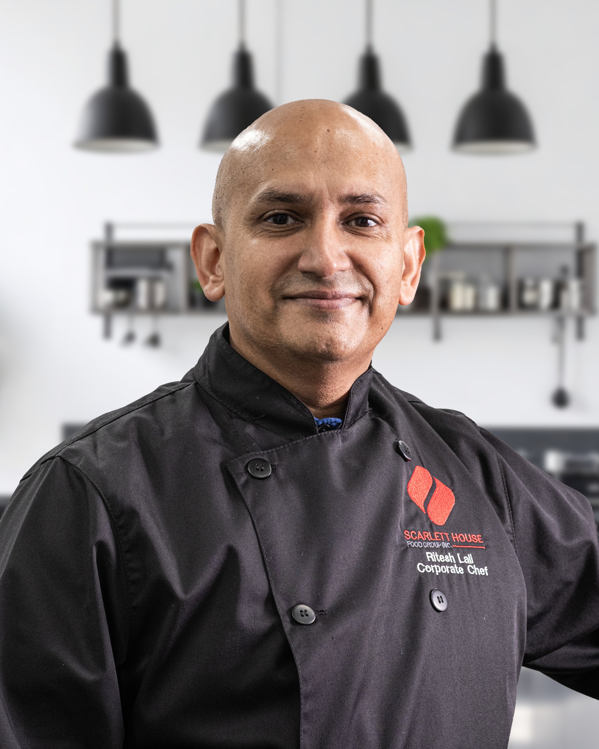 Close-up of a man dressed as a chef in a professional kitchen. He is wearing a black chef's coat with red and white embroidered text identifying him as Ritesh Lal, a corporate chef at Scarlett House Food Group Inc. The background shows modern kitchen lighting and shelving.