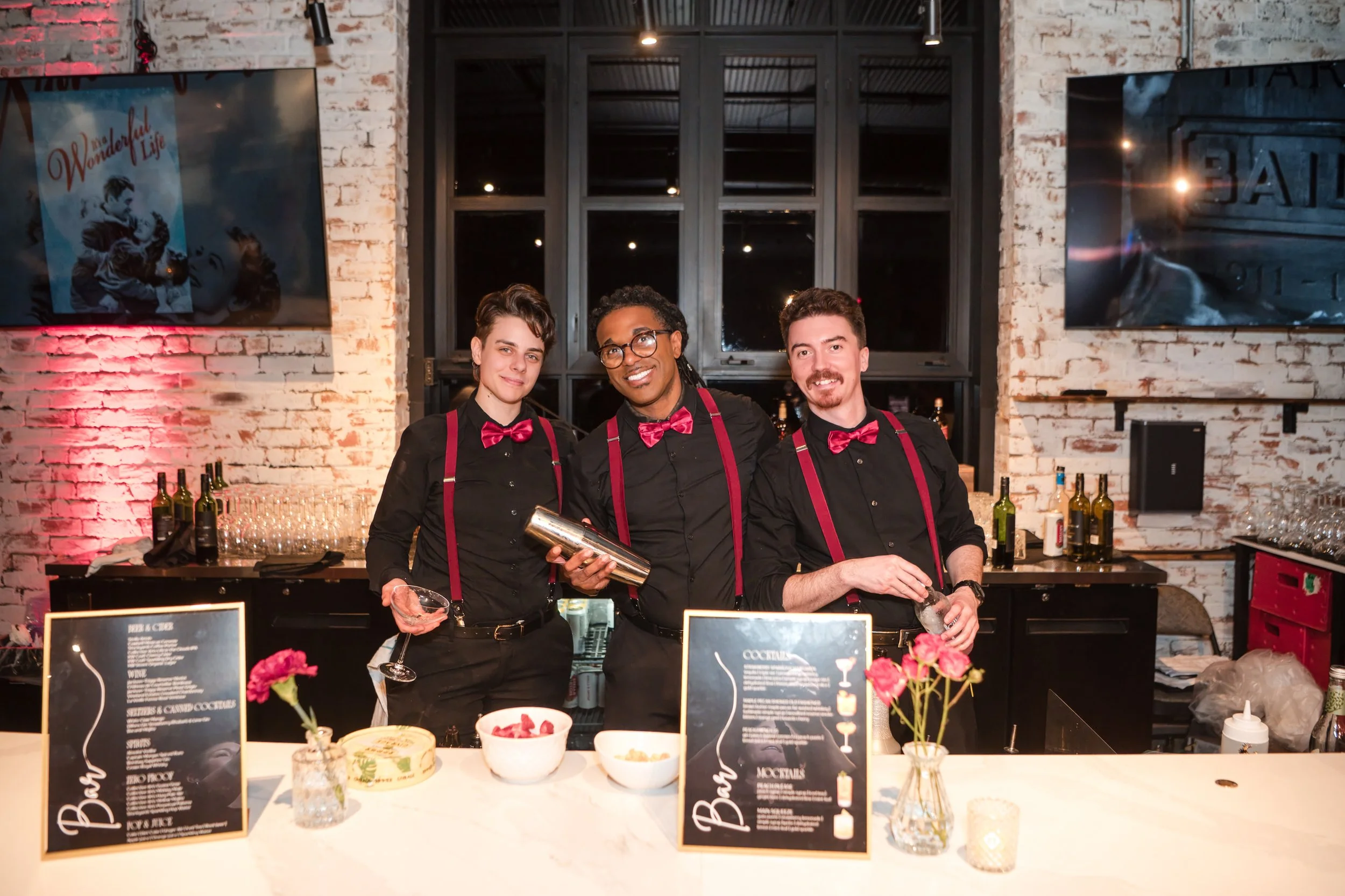 Three smiling bartenders in black shirts with red suspenders and bow ties behind a bar counter with cocktail menus and decorative flowers, in a brick-walled room with large windows and two TV screens.