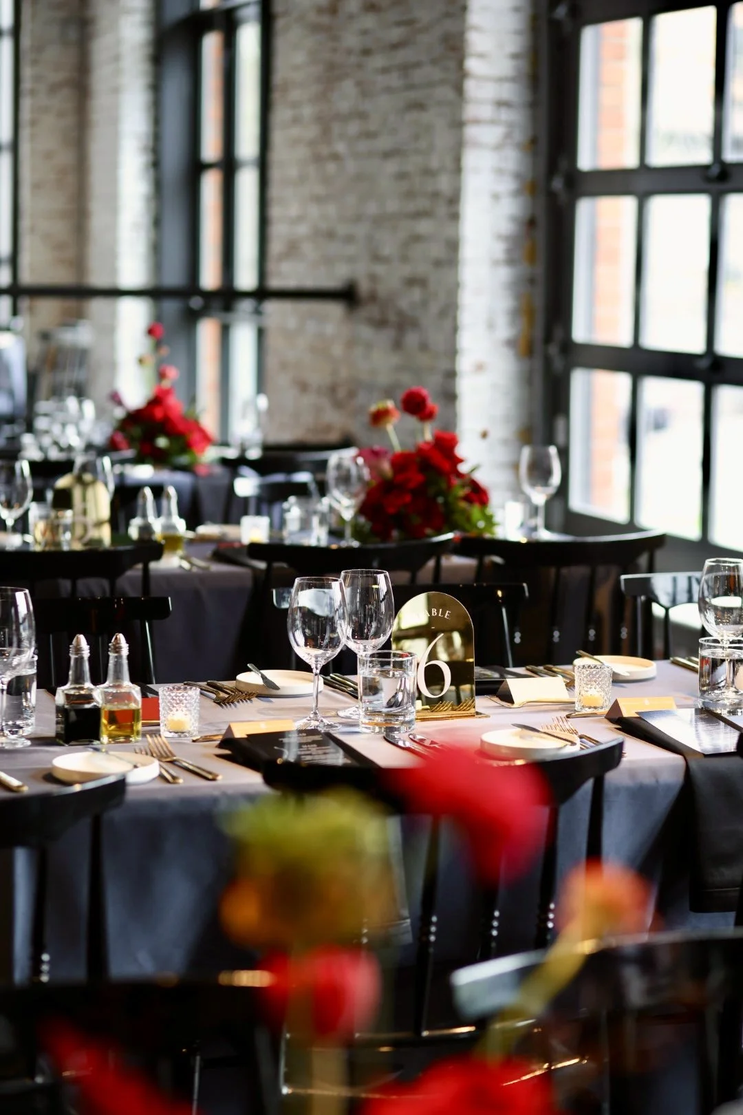 Elegant banquet table setup with glassware, cutlery, floral centerpieces, and table number in a stylish industrial venue with large windows and exposed brick walls.
