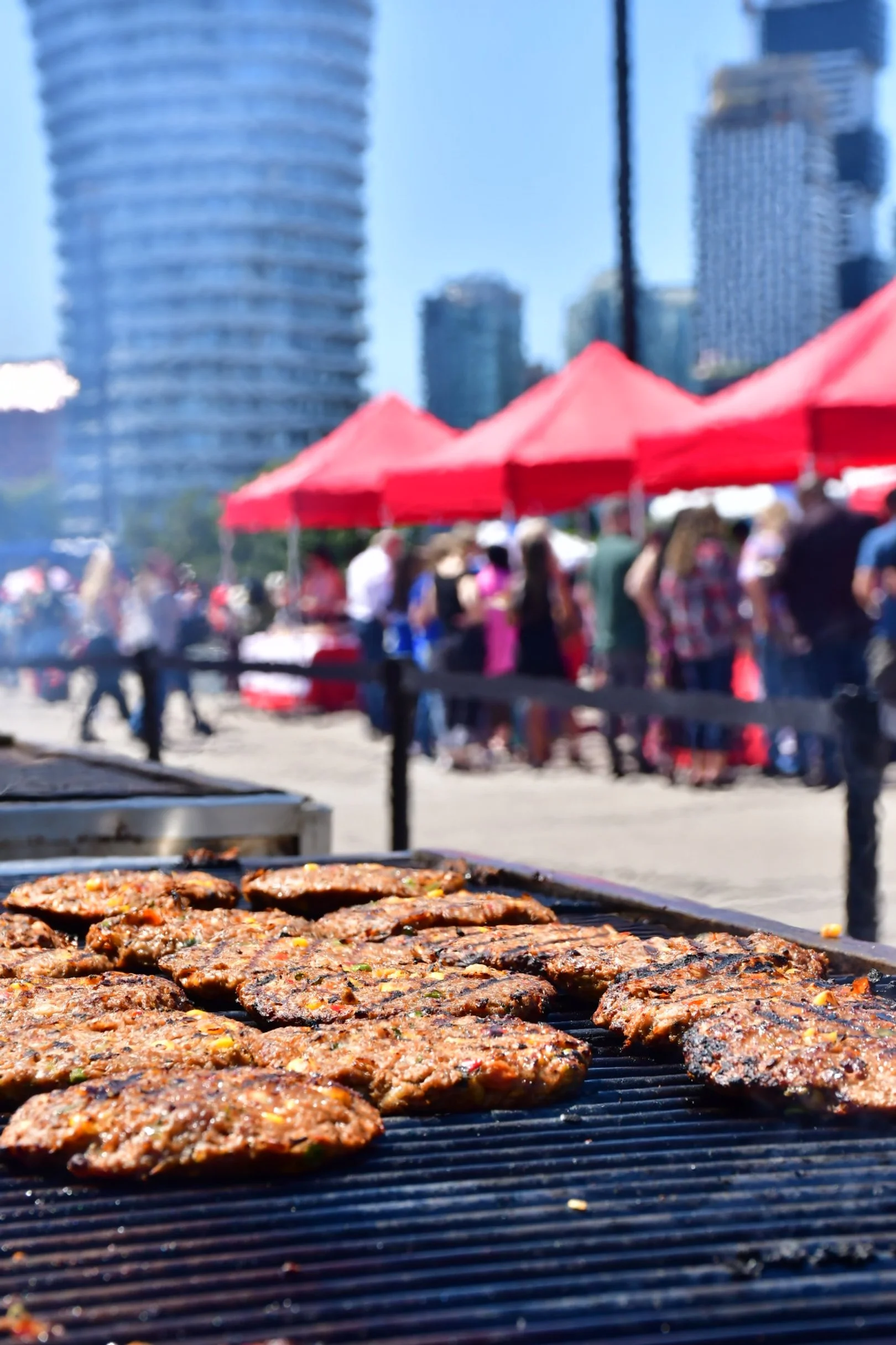 Close-up of grilled veggie burger patties cooking on a barbecue grill with a lively outdoor event and tall buildings in the background.