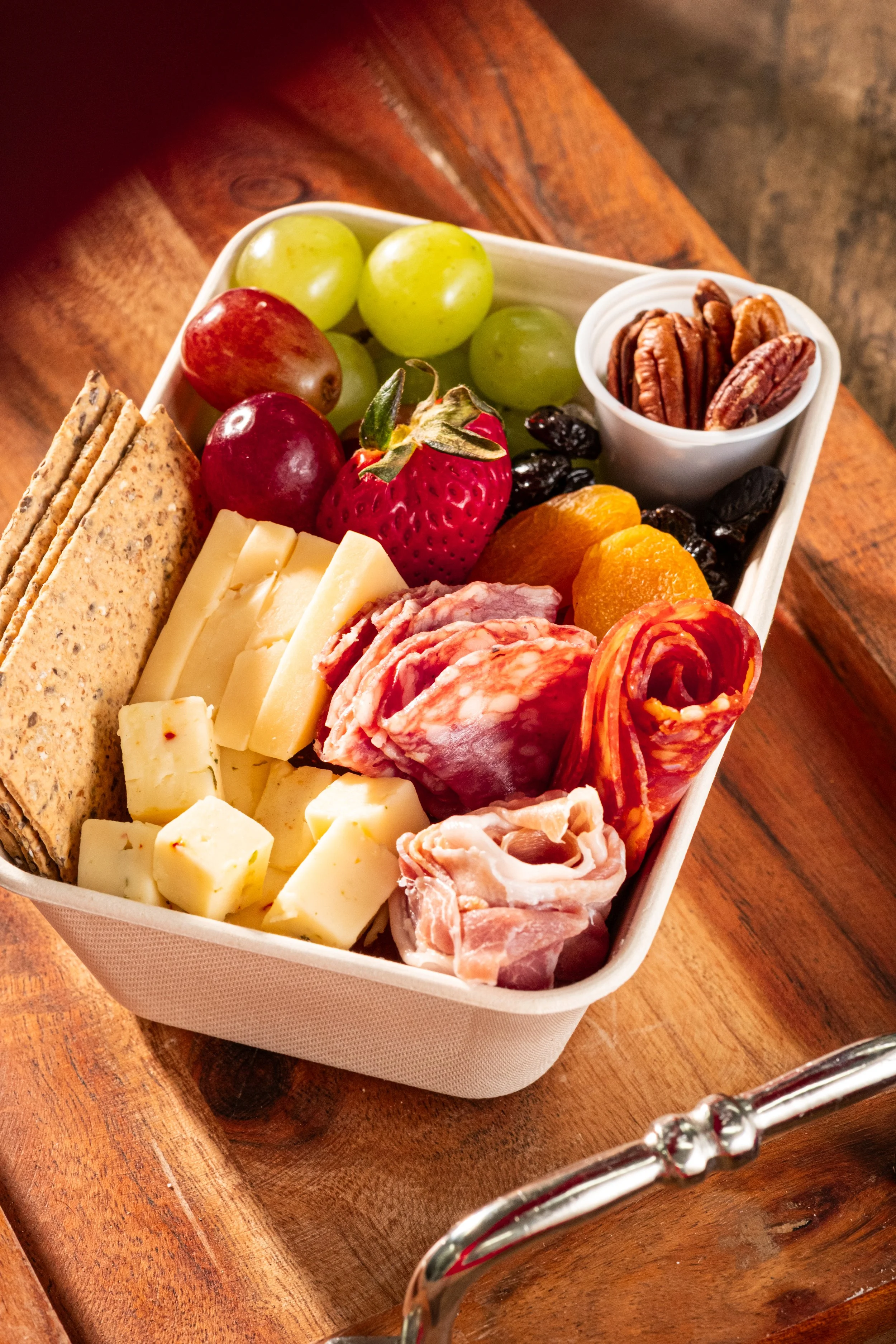 A cheese and charcuterie board with grapes, strawberries, dried apricots, pecans, crackers, various cheeses, salami, and prosciutto on a wooden surface.