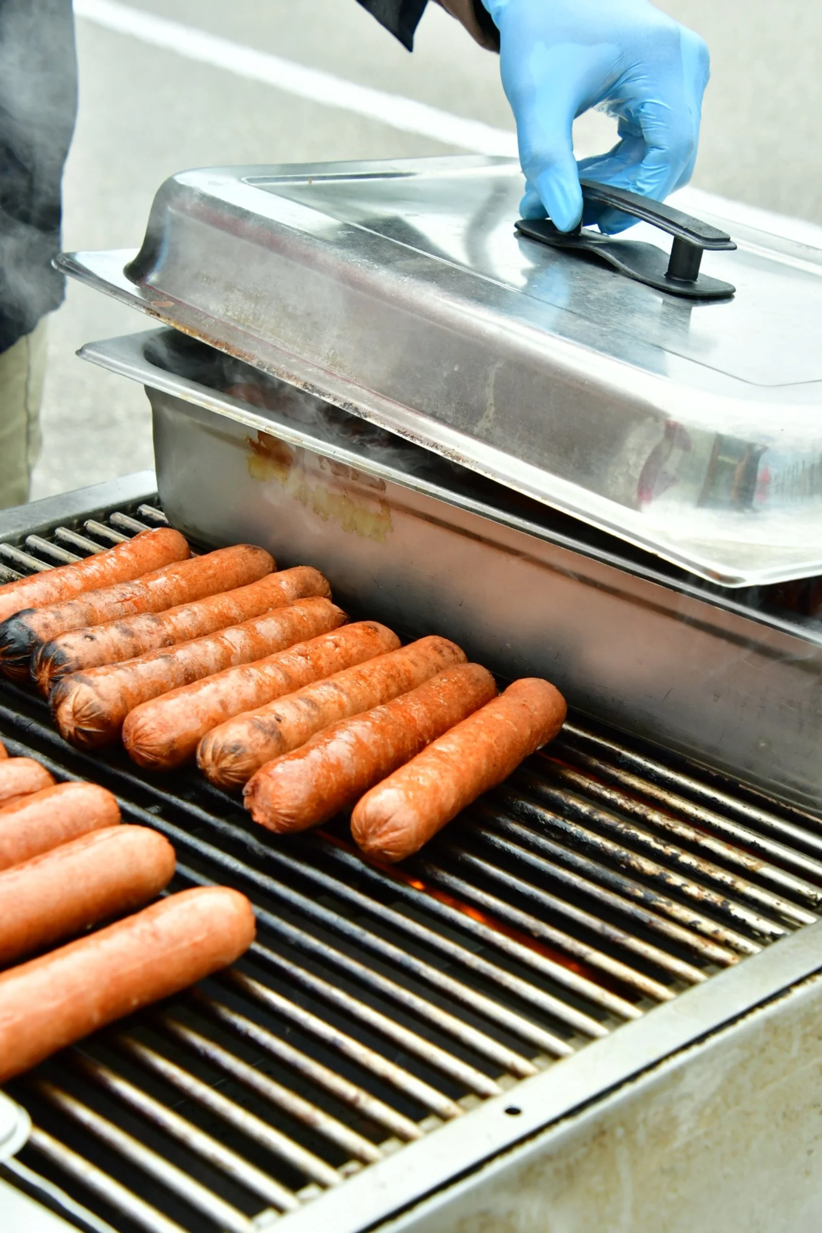 Hot dogs being grilled on a barbecue grill with steam rising and a person lifting a metal cover with a gloved hand.