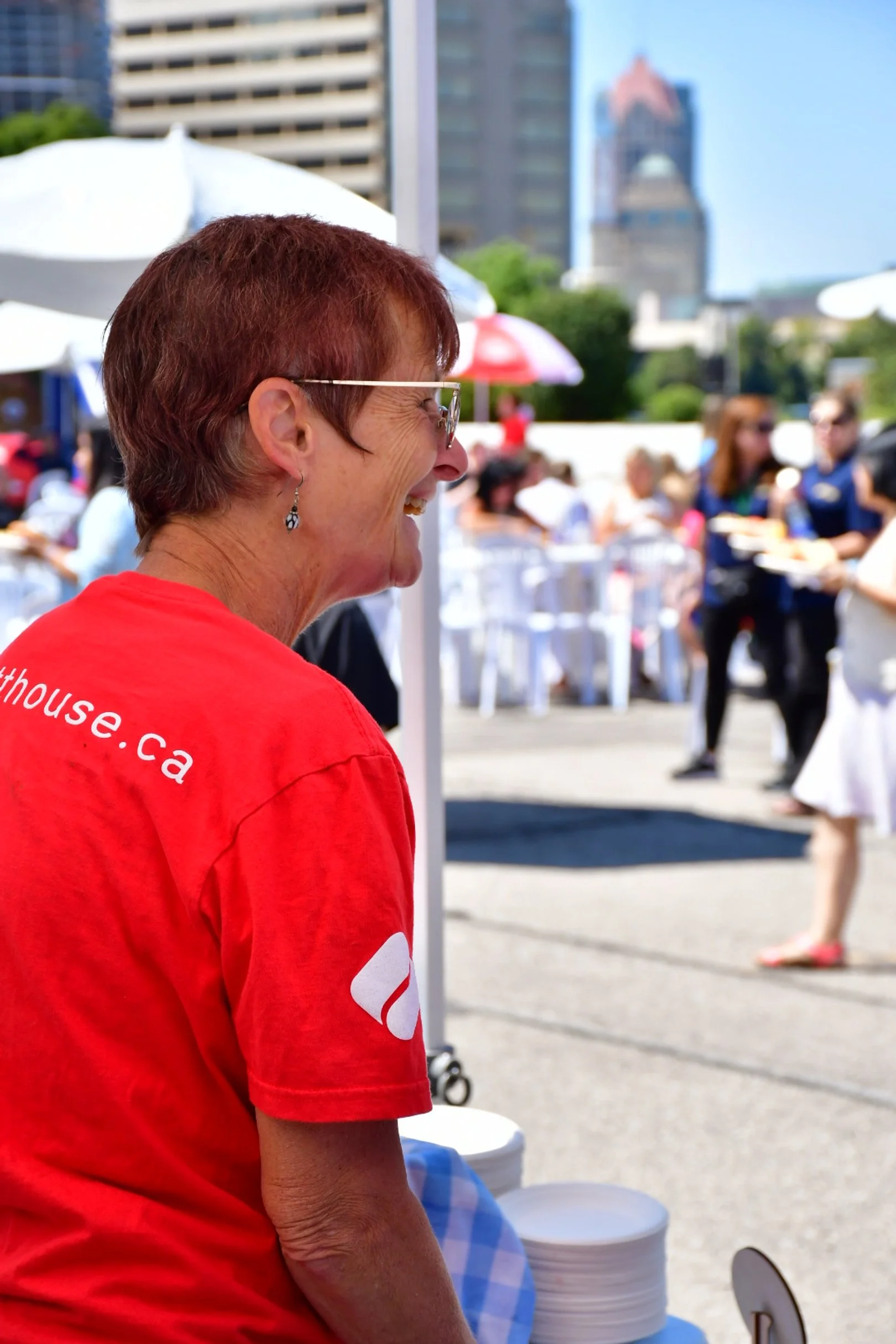Woman in a red shirt smiling at an outdoor event, with tables and other people in the background.