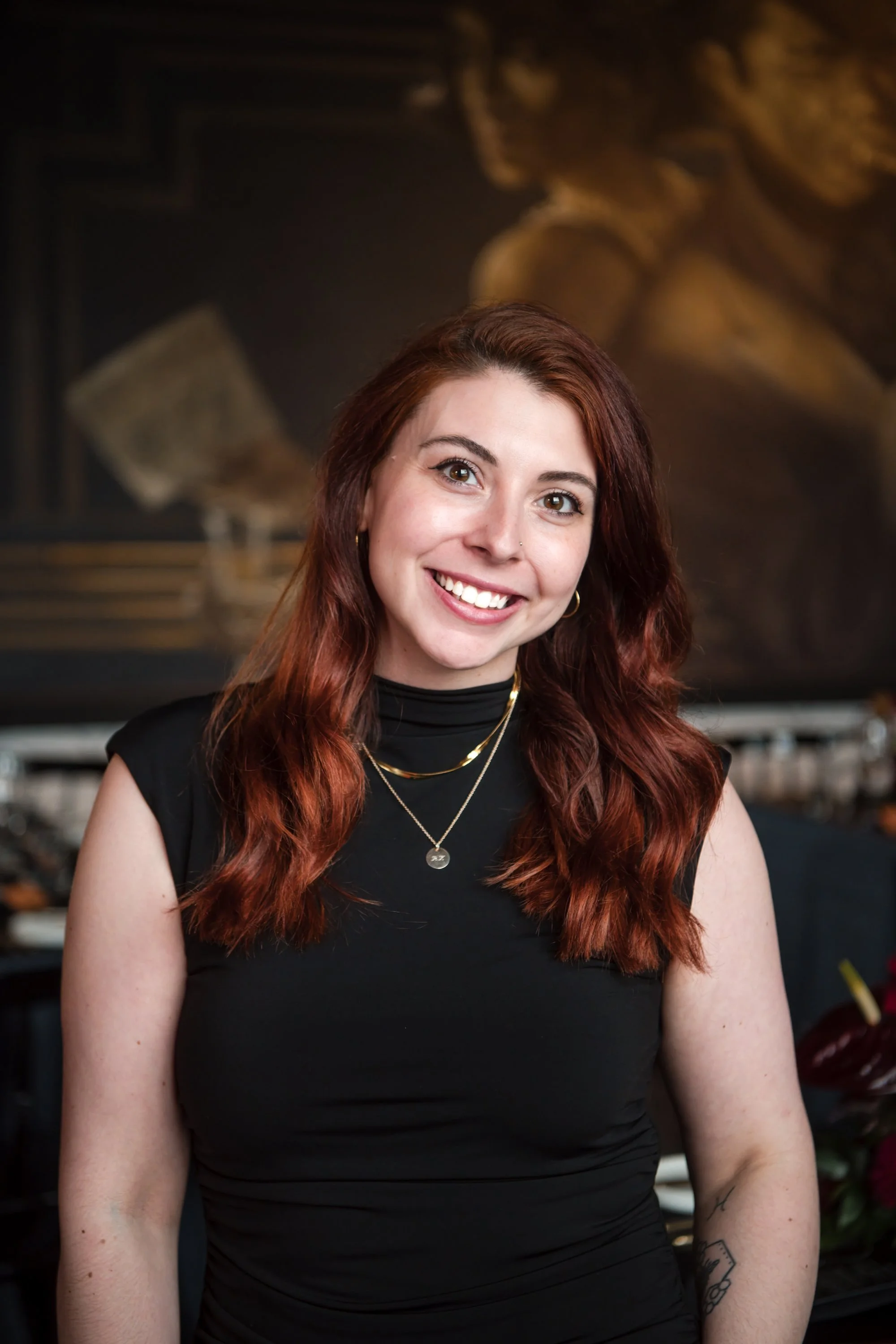 A portrait of a young woman with long, wavy auburn hair smiling in front of a dark, artistic background. This is Caiti, the catering & events assistant.