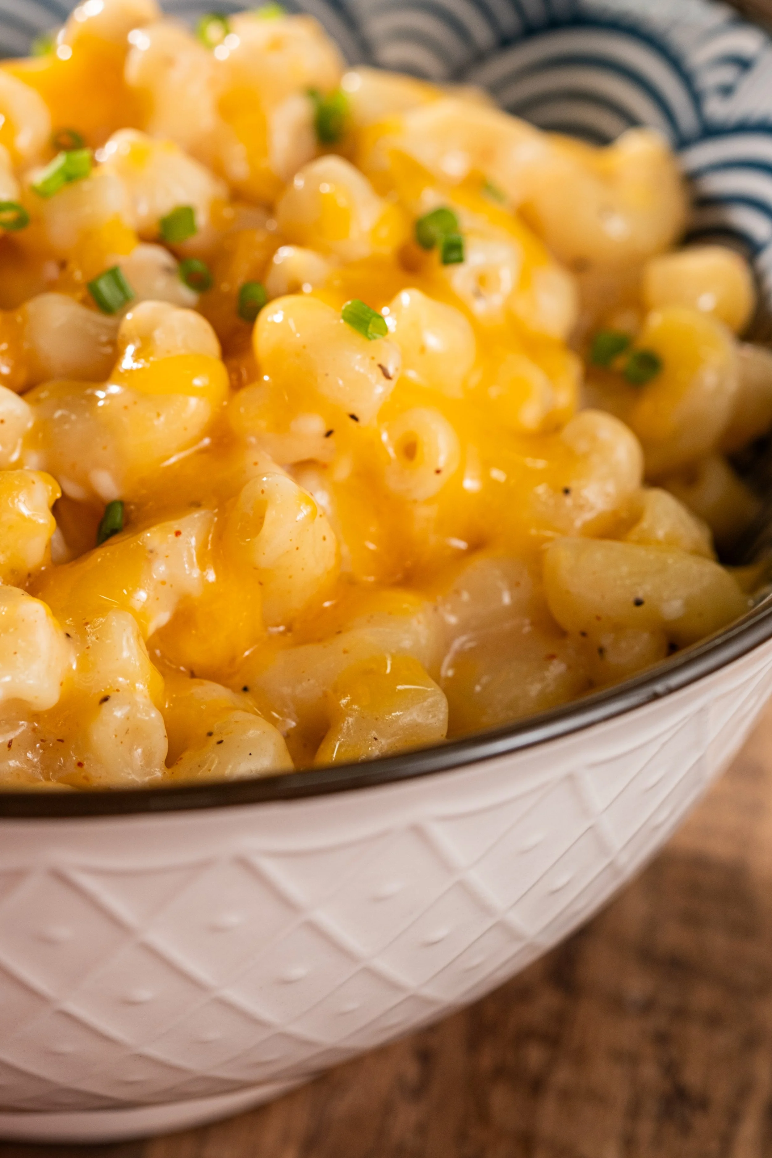 Close-up of creamy macaroni and cheese with shredded cheddar cheese topping, garnished with chopped green chives in a white textured bowl on a wooden surface.