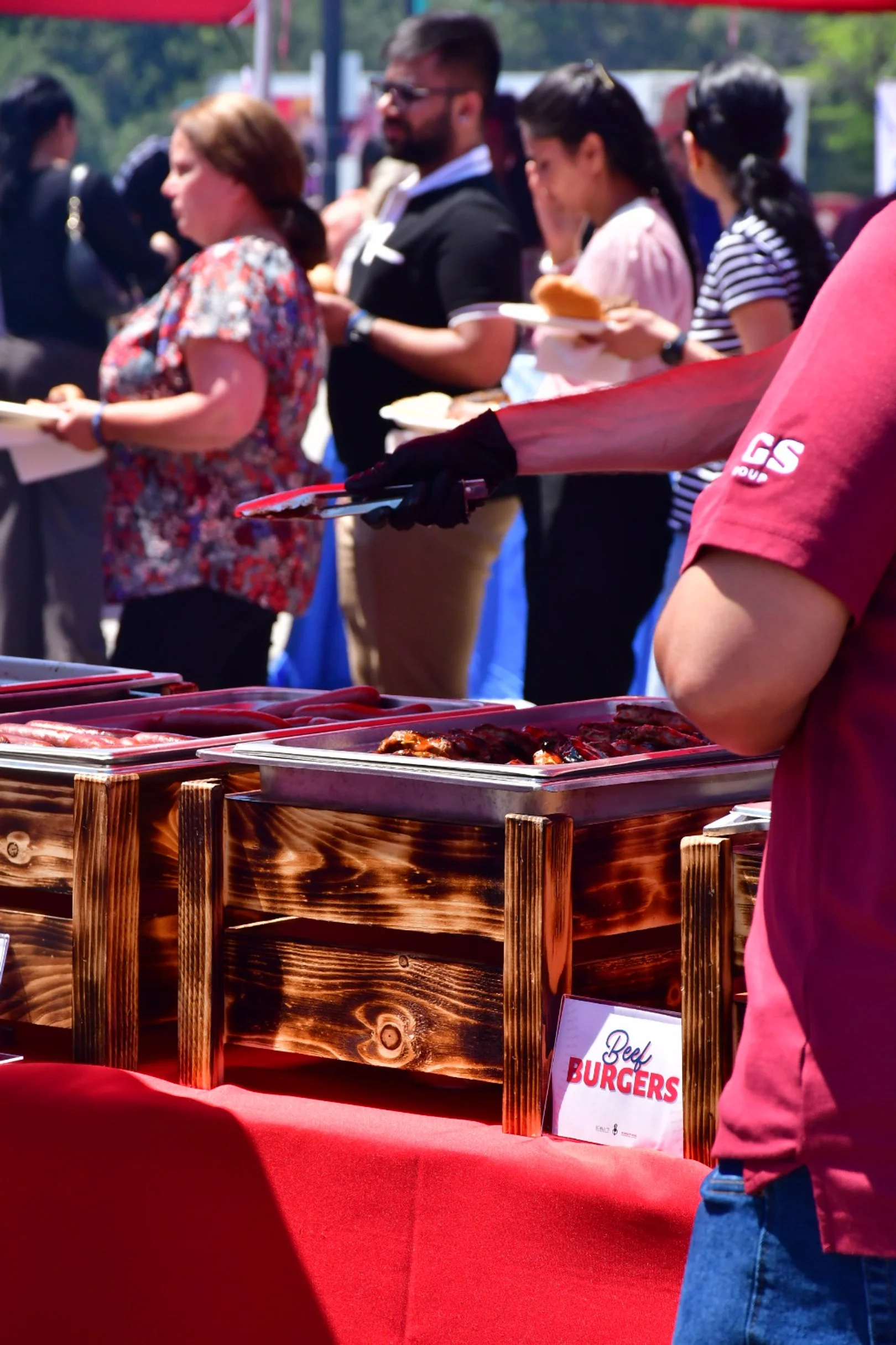 People standing in line at a food buffet, with a focus on a person serving barbecue food from a tray, at an outdoor event on a sunny day.