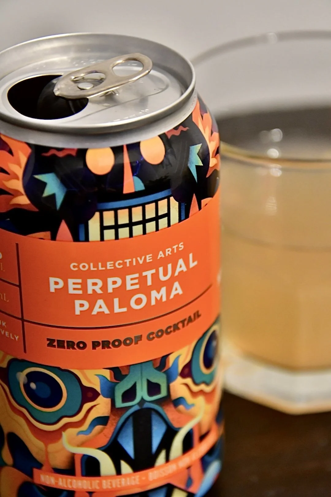 A colorful can of Collective Arts Perpetual Paloma zero-proof cocktail with a geometric, Day of the Dead-inspired design, sitting next to a glass of pale beverage.
