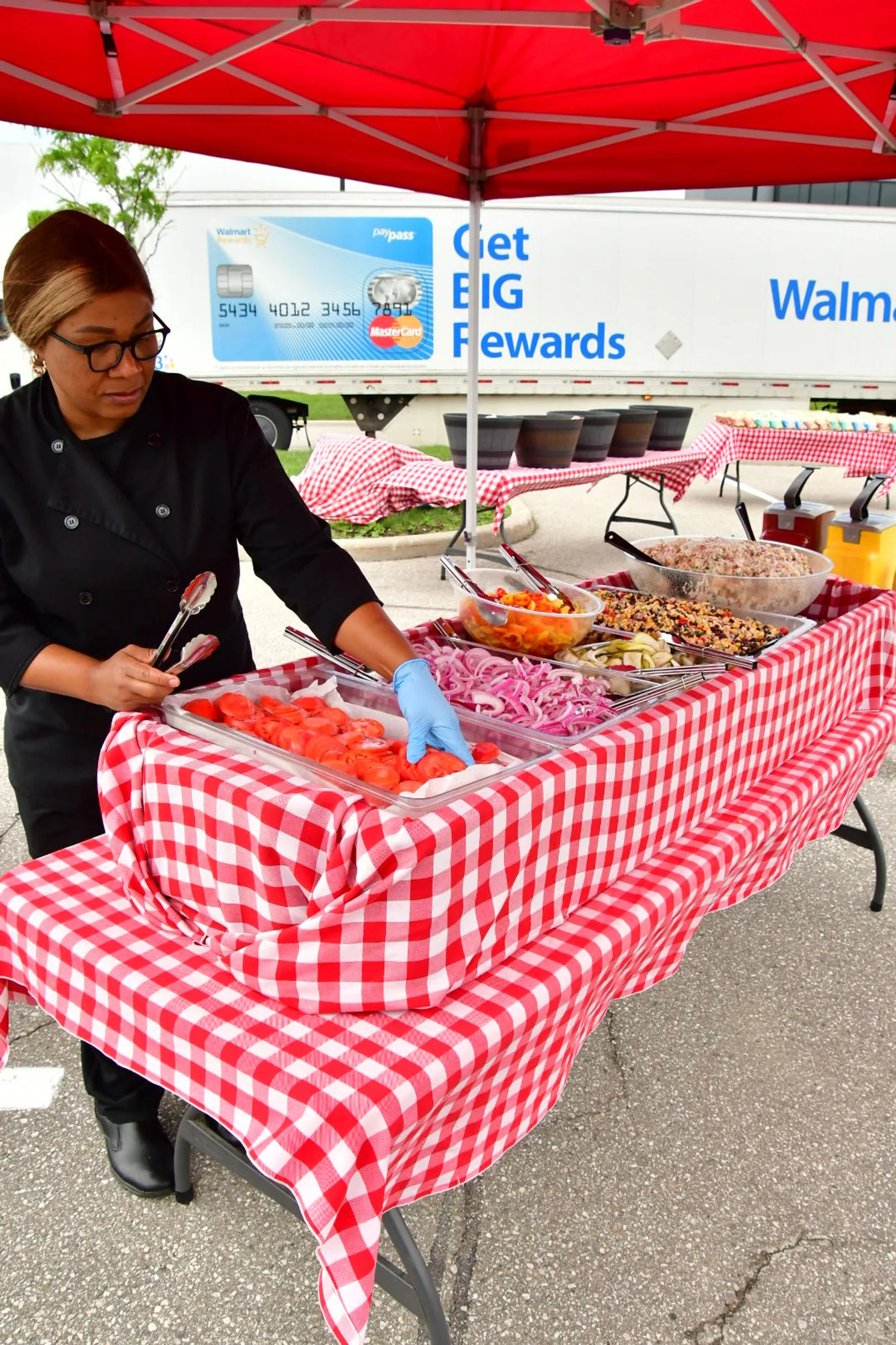 A woman in a black chef's uniform and glasses prepares food at a picnic-style buffet table covered with a red and white checkered tablecloth, with various salads and toppings, under a red canopy outdoors.