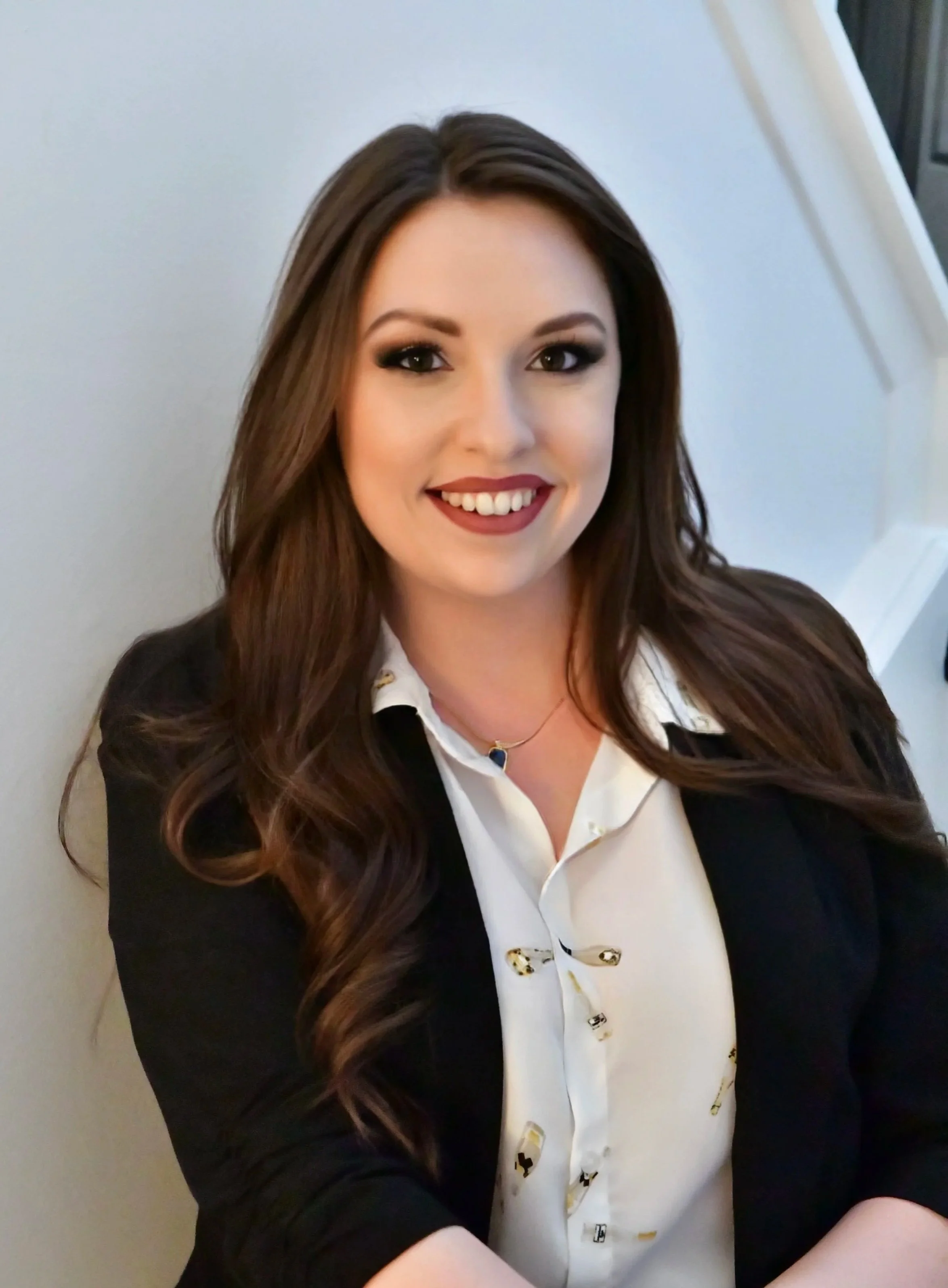 A woman with long, wavy brown hair and makeup, wearing a white top and a black blazer, smiling at the camera.