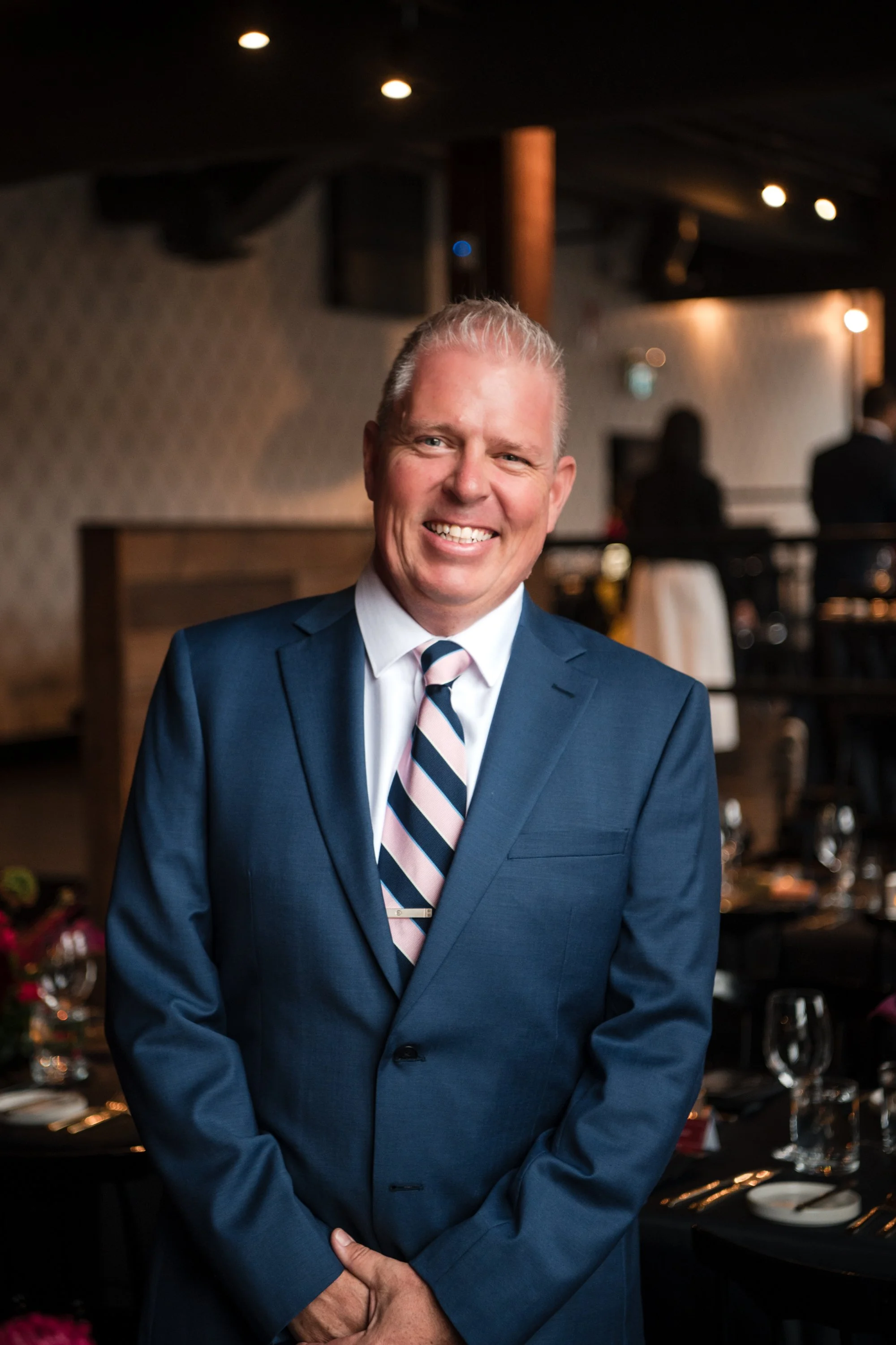 Man in blue suit, white shirt, and striped tie smiling and posing indoors at a formal event.