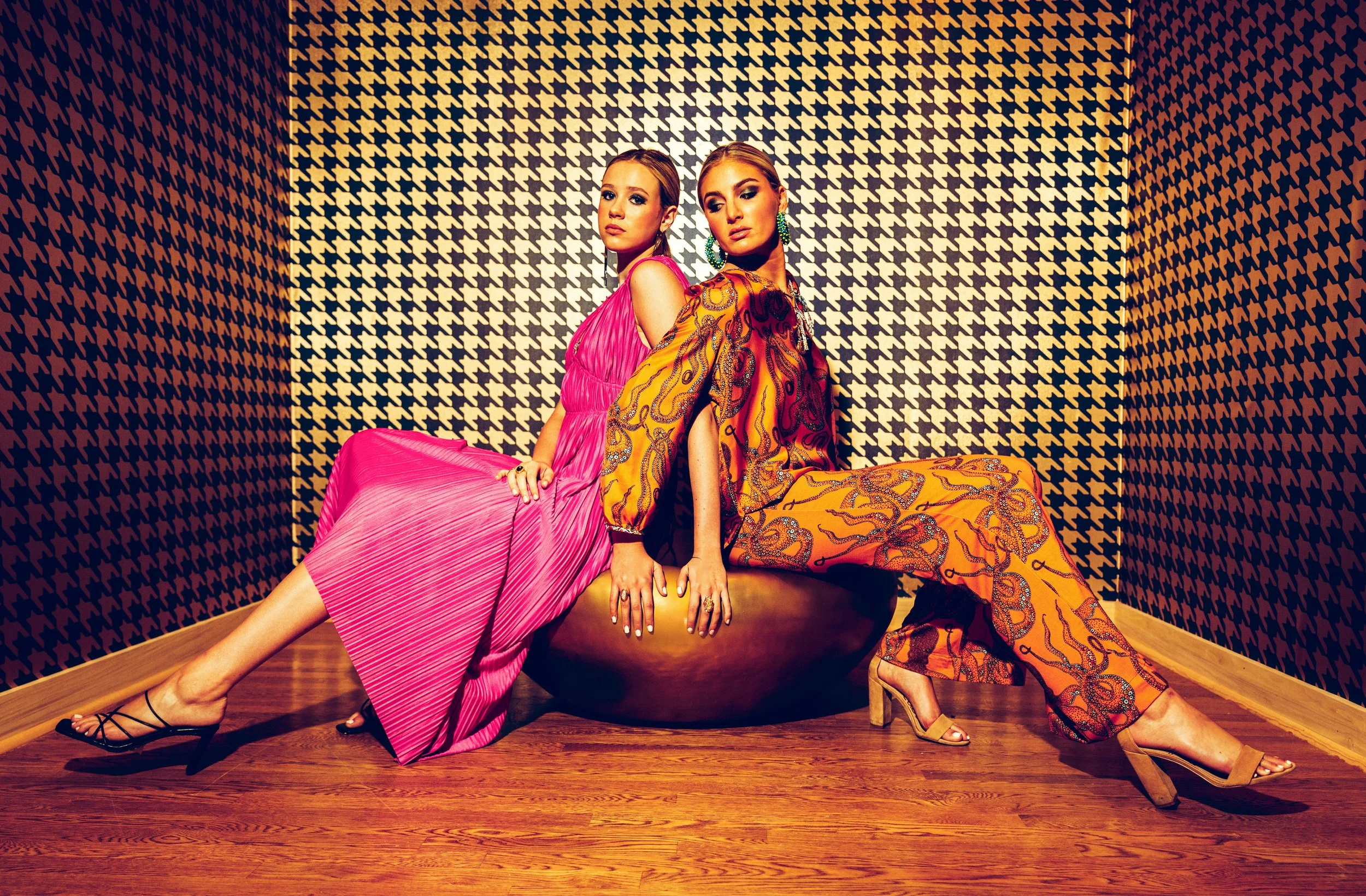Two fashionably dressed women sitting on a large golden bowl in a room with black and beige houndstooth patterned walls and wooden floors.