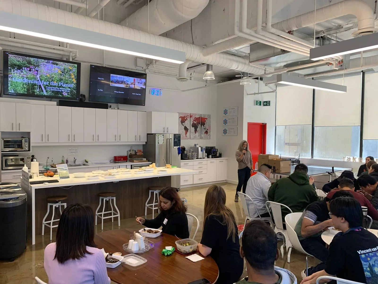 People sitting at tables and eating in a cafeteria with a kitchen counter, microwave, and TV screens on the wall.