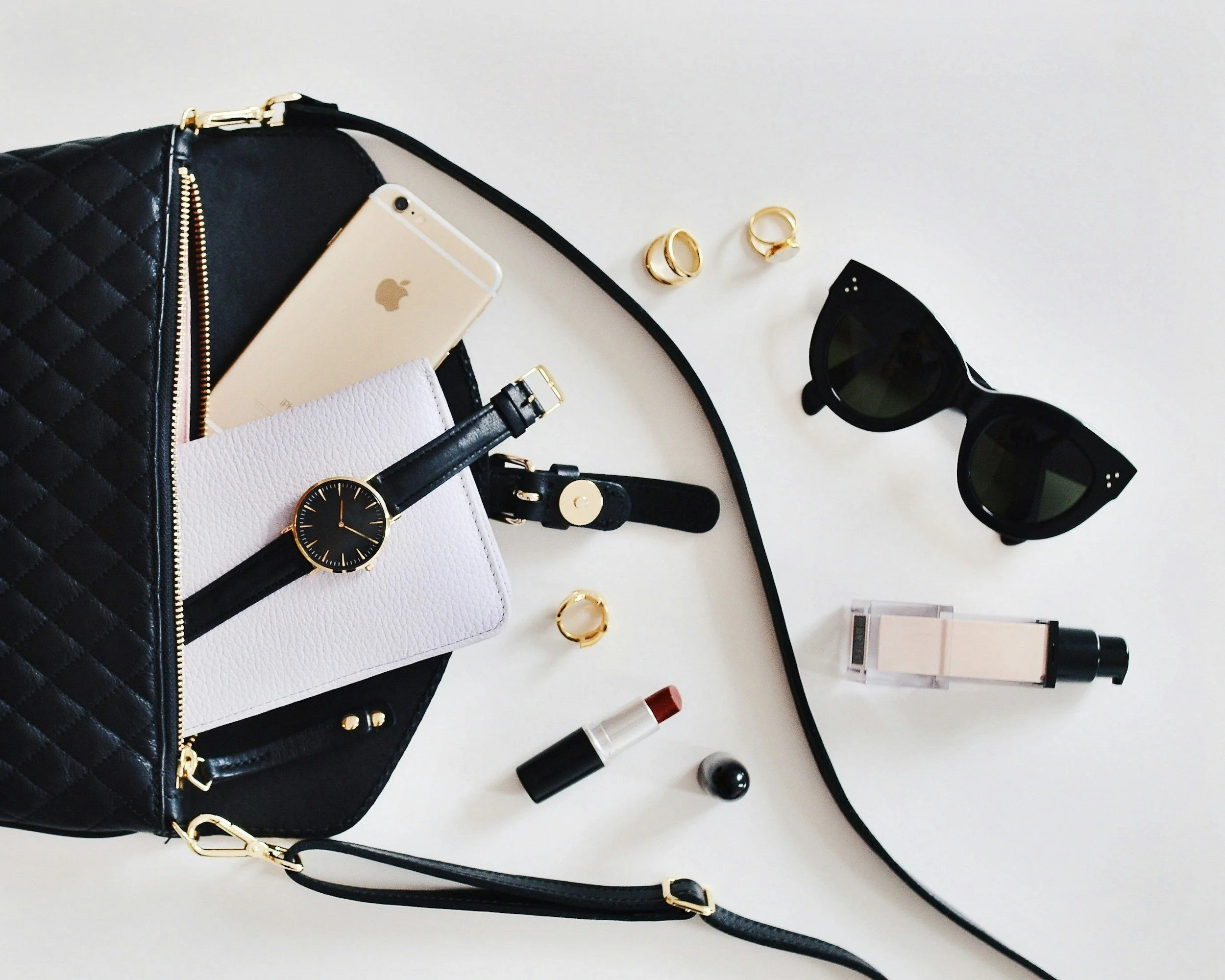 Black quilted handbag spilling over with a gold iPhone, white wallet, black watch, and gold jewelry, alongside black sunglasses, perfume, and makeup on a white surface.