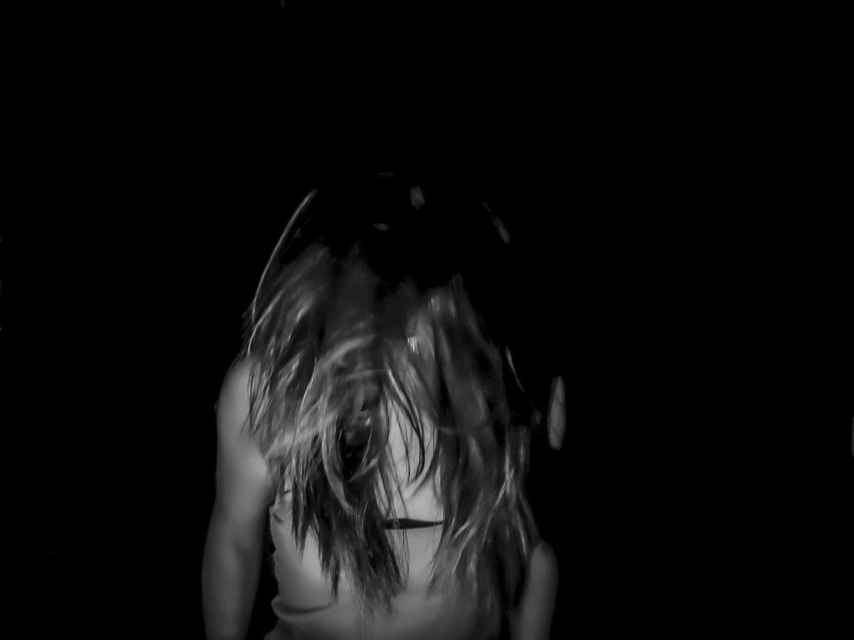 A person with long, wavy hair wearing a light-colored tank top standing in a dark space, head tilted downward, with the hair covering the face.