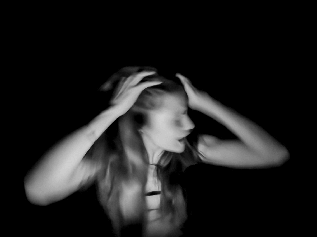 Black and white blurred photo of a woman with long hair, holding her head with both hands, against a dark background.