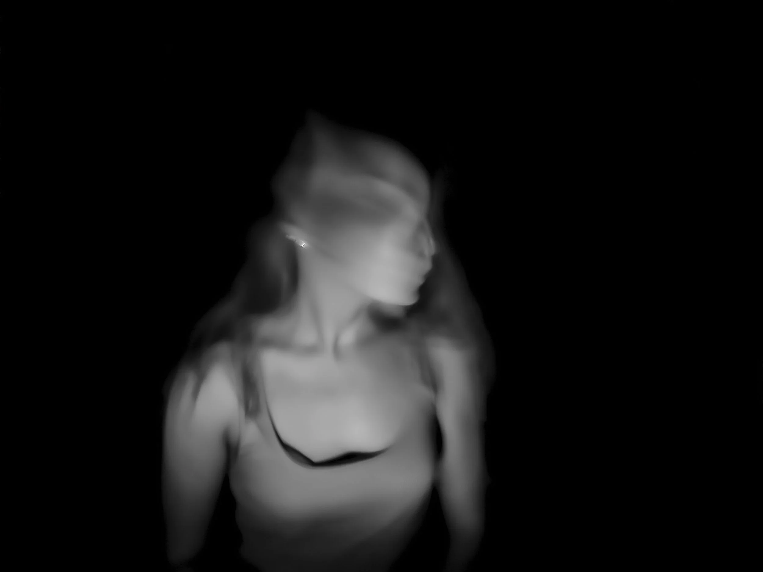 Black and white, blurred photo of a woman with long hair, wearing a spaghetti strap top, looking to the side against a dark background.