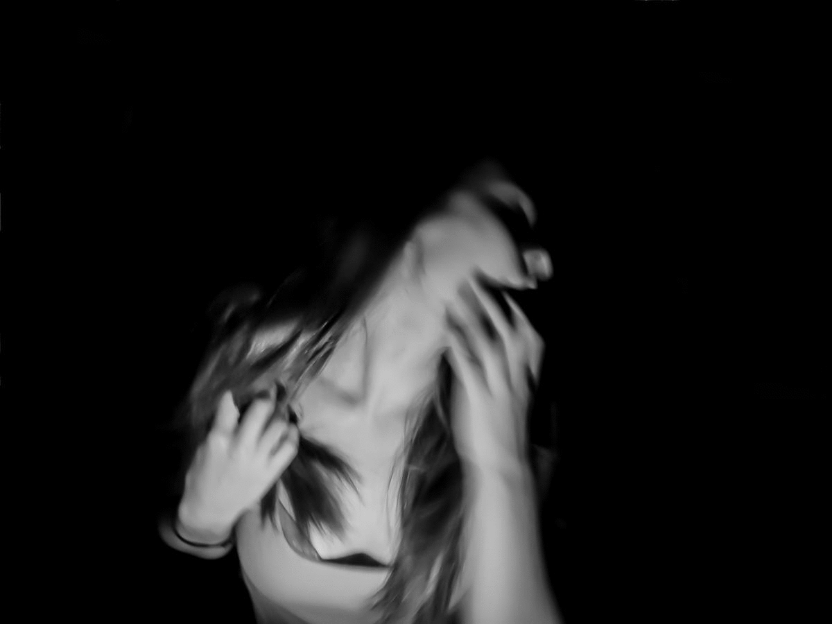 Blurred black and white photo of a woman with long hair, touching her face with one hand and her hair with the other, against a dark background.