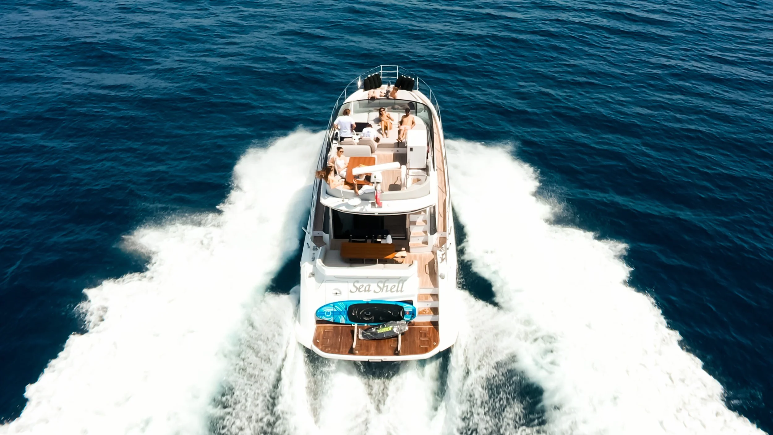 A white yacht named Sea Shell cruising on blue ocean water with five people on board.