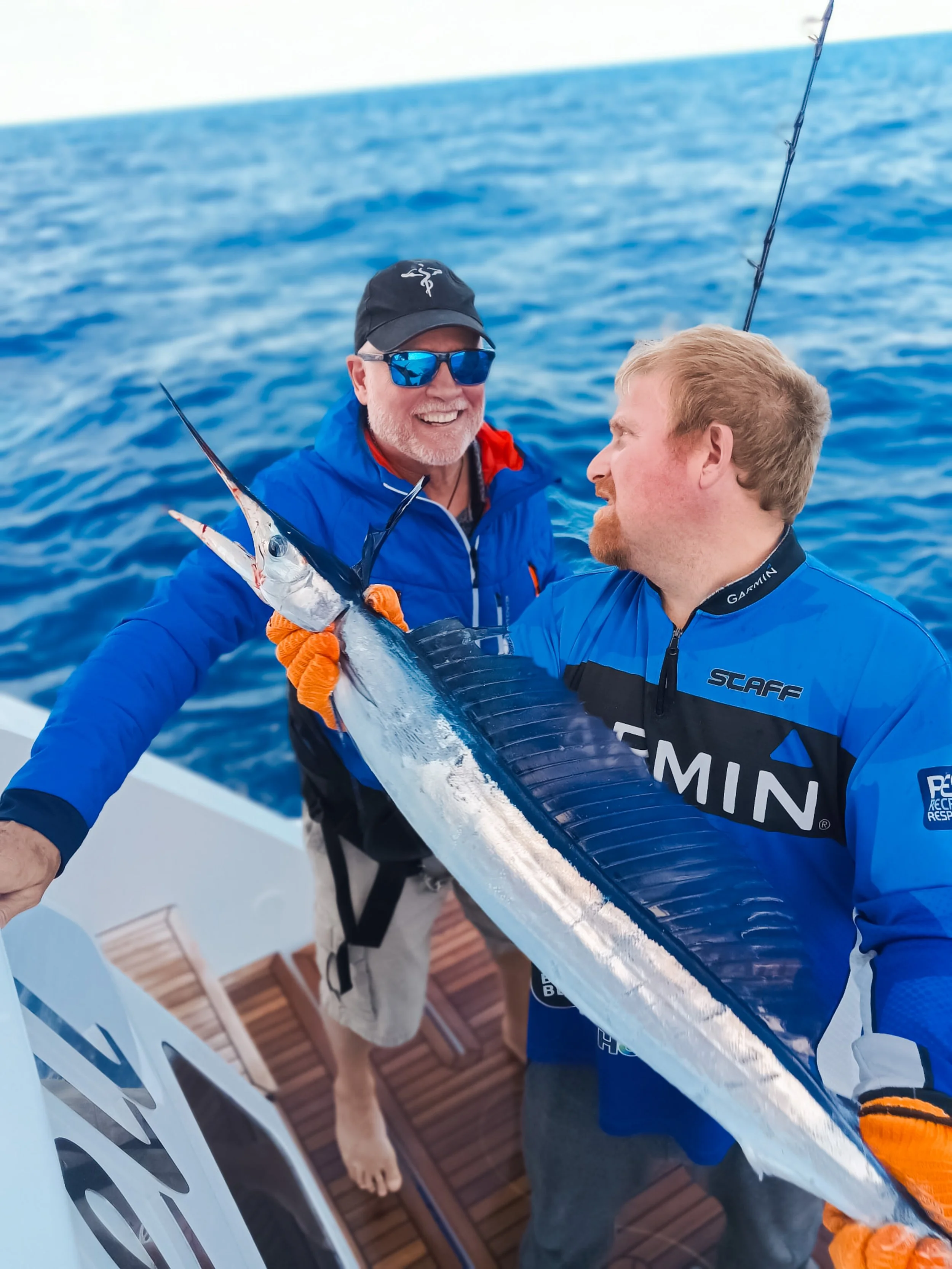 Two men on a boat holding a large swordfish after a successful catch. One man is smiling and wearing sunglasses, a black cap, and a blue jacket, while the other is looking at the fish and wearing a blue and black fishing jersey. The ocean is in the b