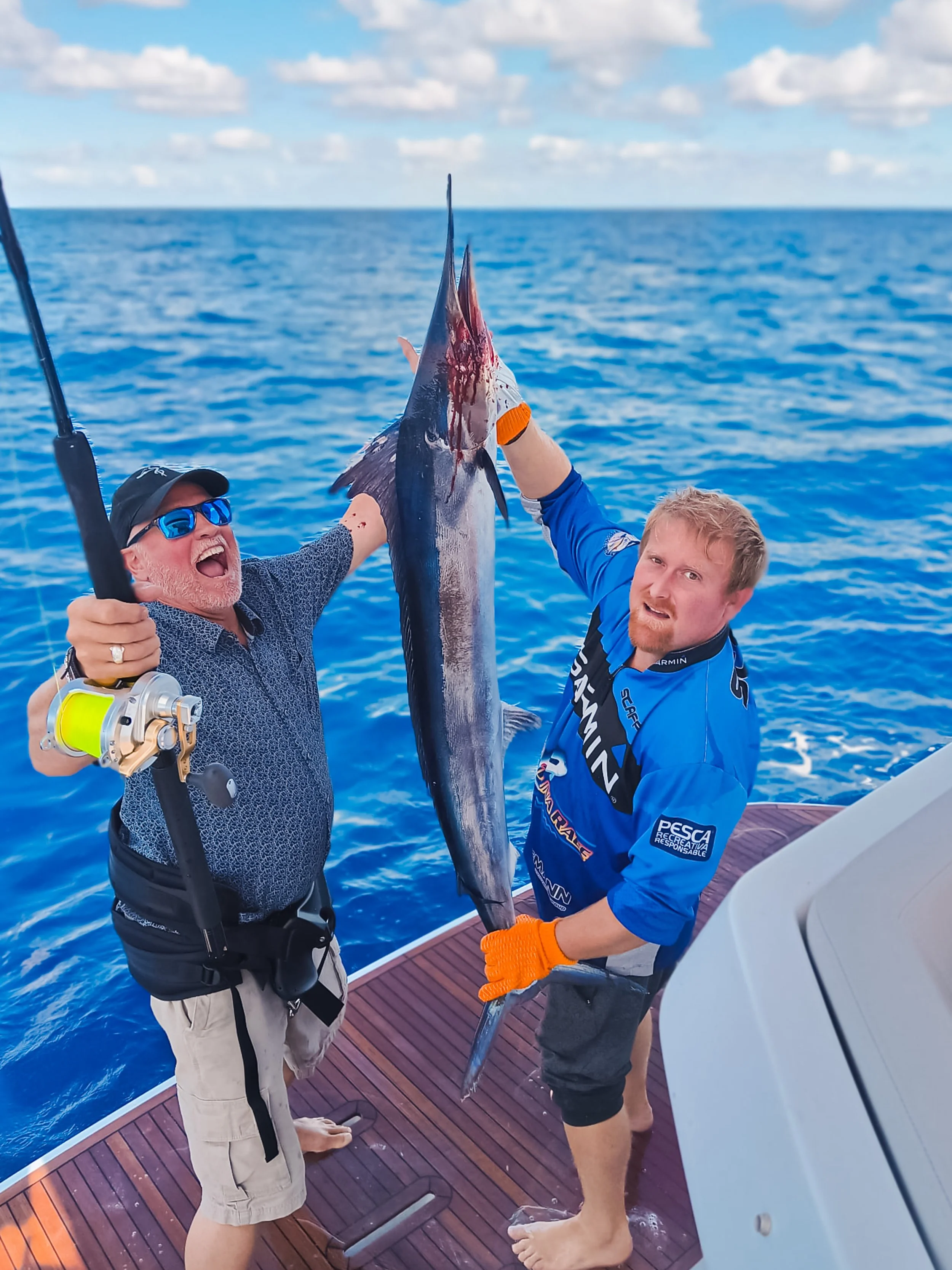 Two men on a boat holding a large fish, with the ocean and sky in the background.