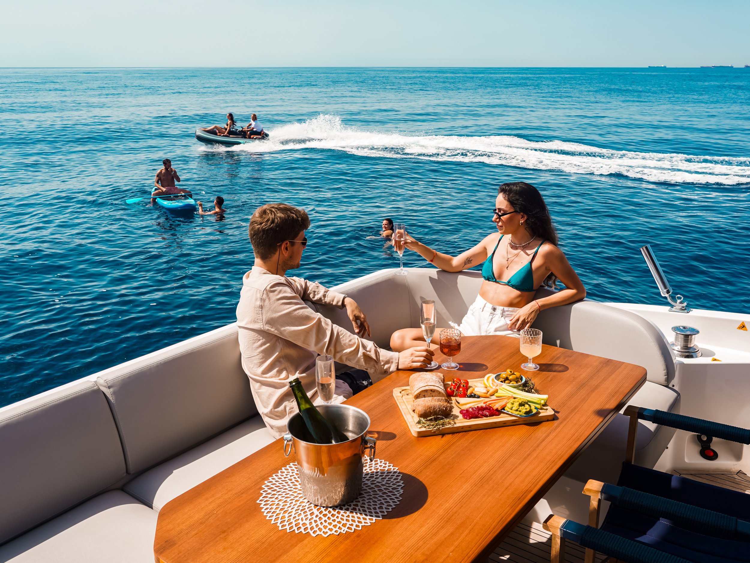 A couple enjoying drinks and food on a yacht with a view of the ocean, with people waterskiing and paddleboarding in the background.