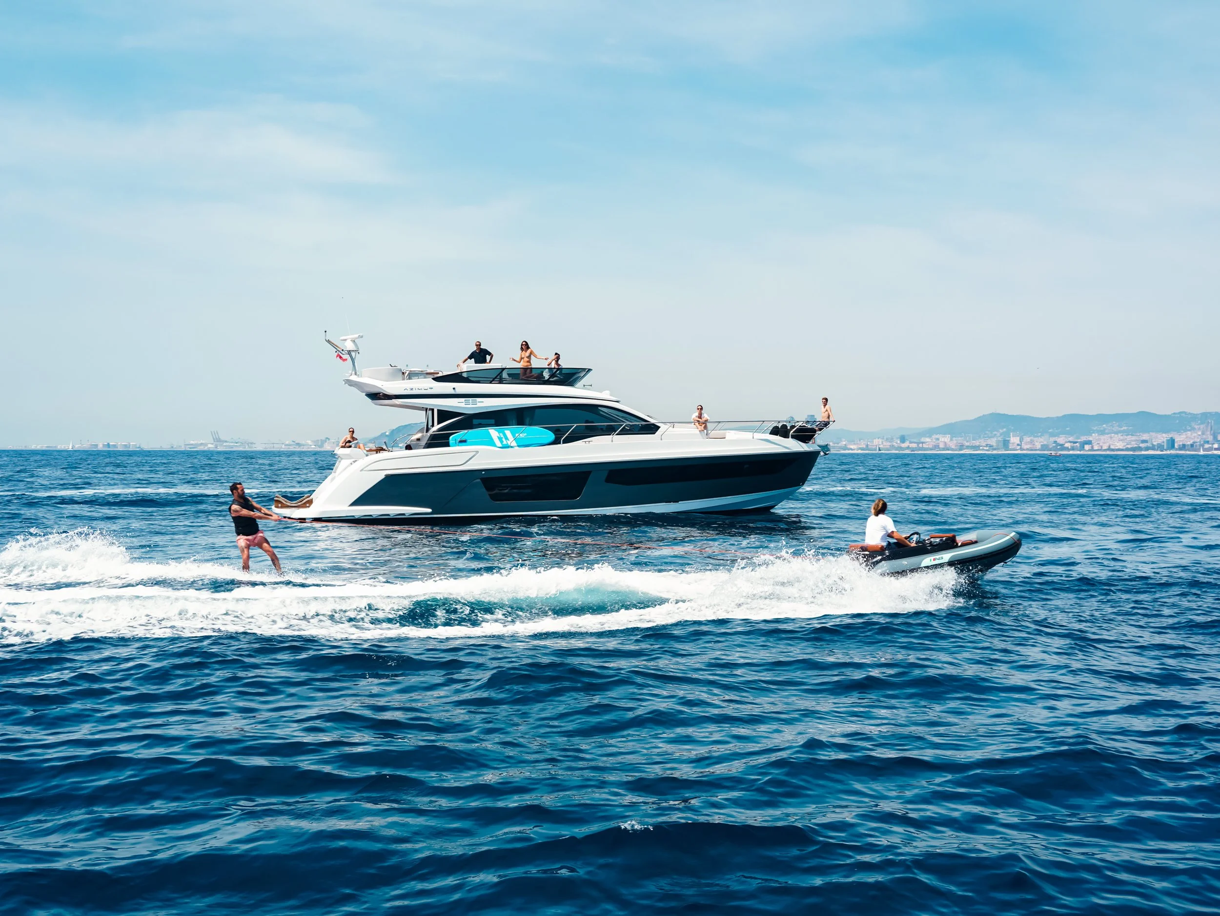A group of people enjoying a day on a yacht and a speedboat on a calm ocean, with a city skyline and mountains in the distance under a partly cloudy sky.