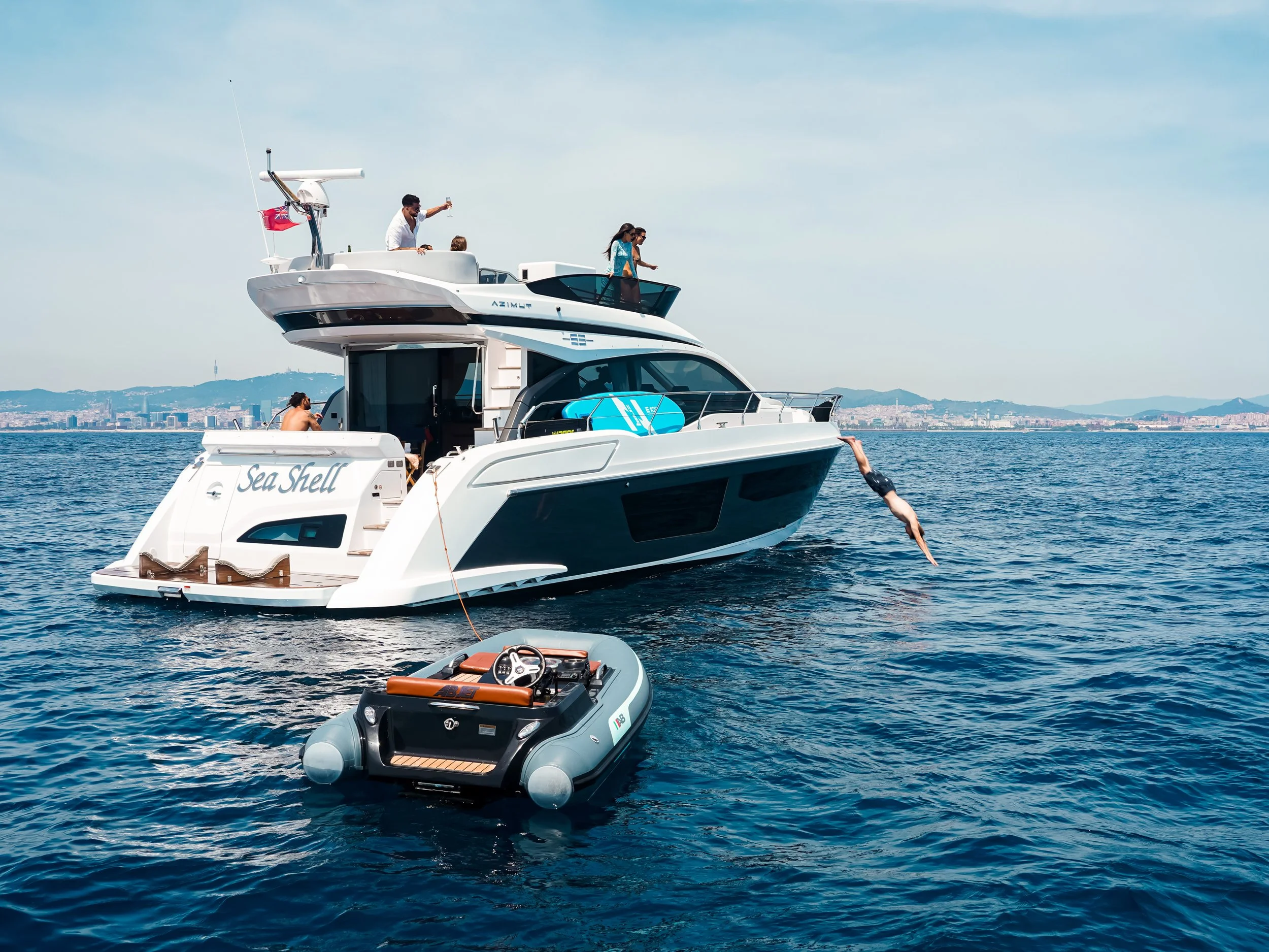 A boat named Sea Shell on the water with a person diving from the side into the water, and several people on the upper deck enjoying the day.
