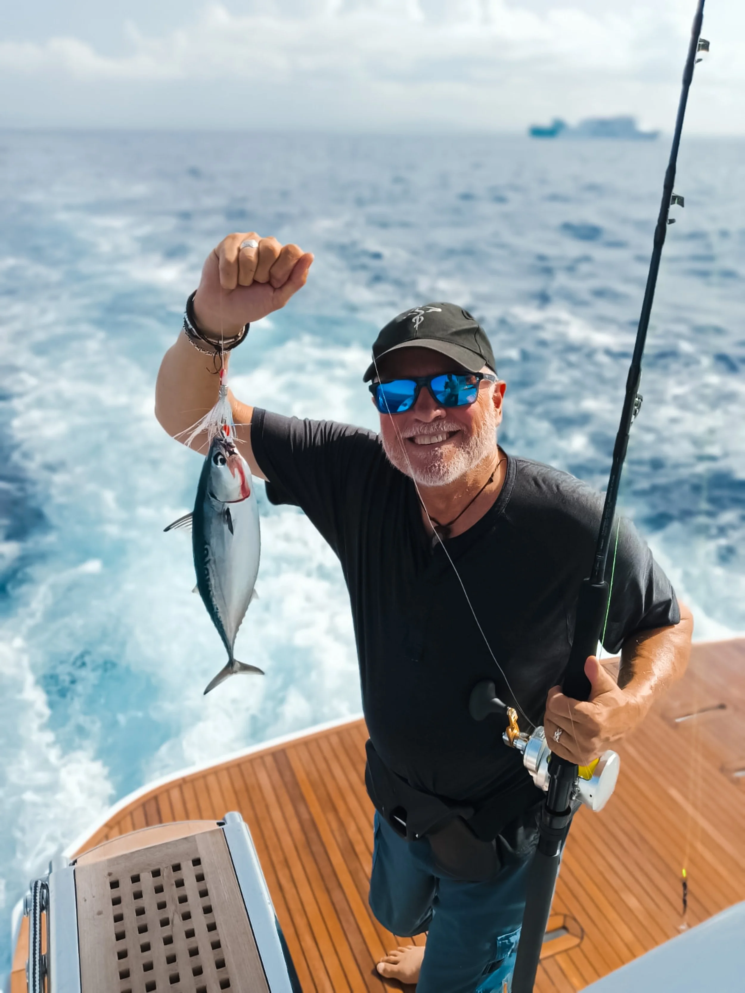 Smiling man in sunglasses and cap holding a fishing rod with a caught fish on a boat, ocean in the background.