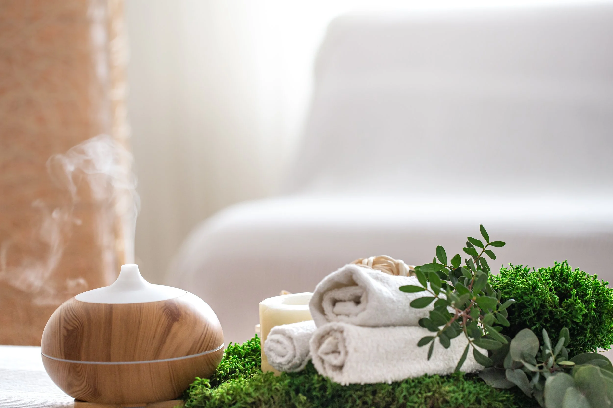 Rolled white towels, green foliage, candles, and a wooden diffuser on a white surface in a relaxing spa setting.