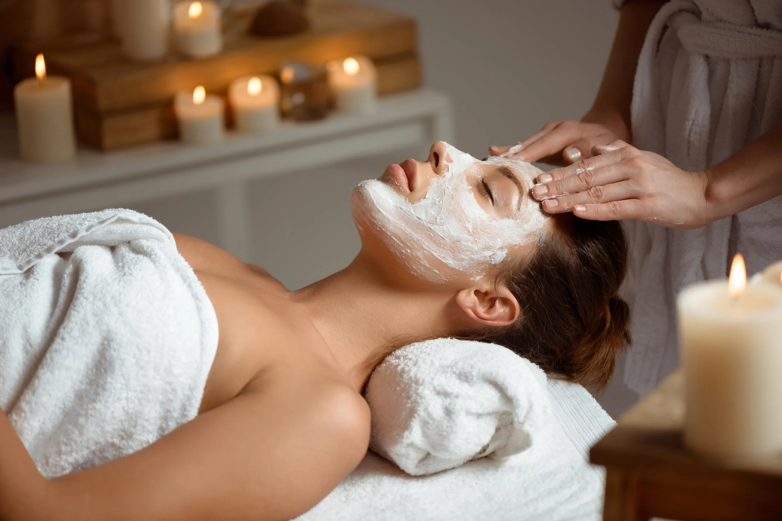 A woman receiving a facial treatment while lying on a massage table in a spa with lit candles in the background.