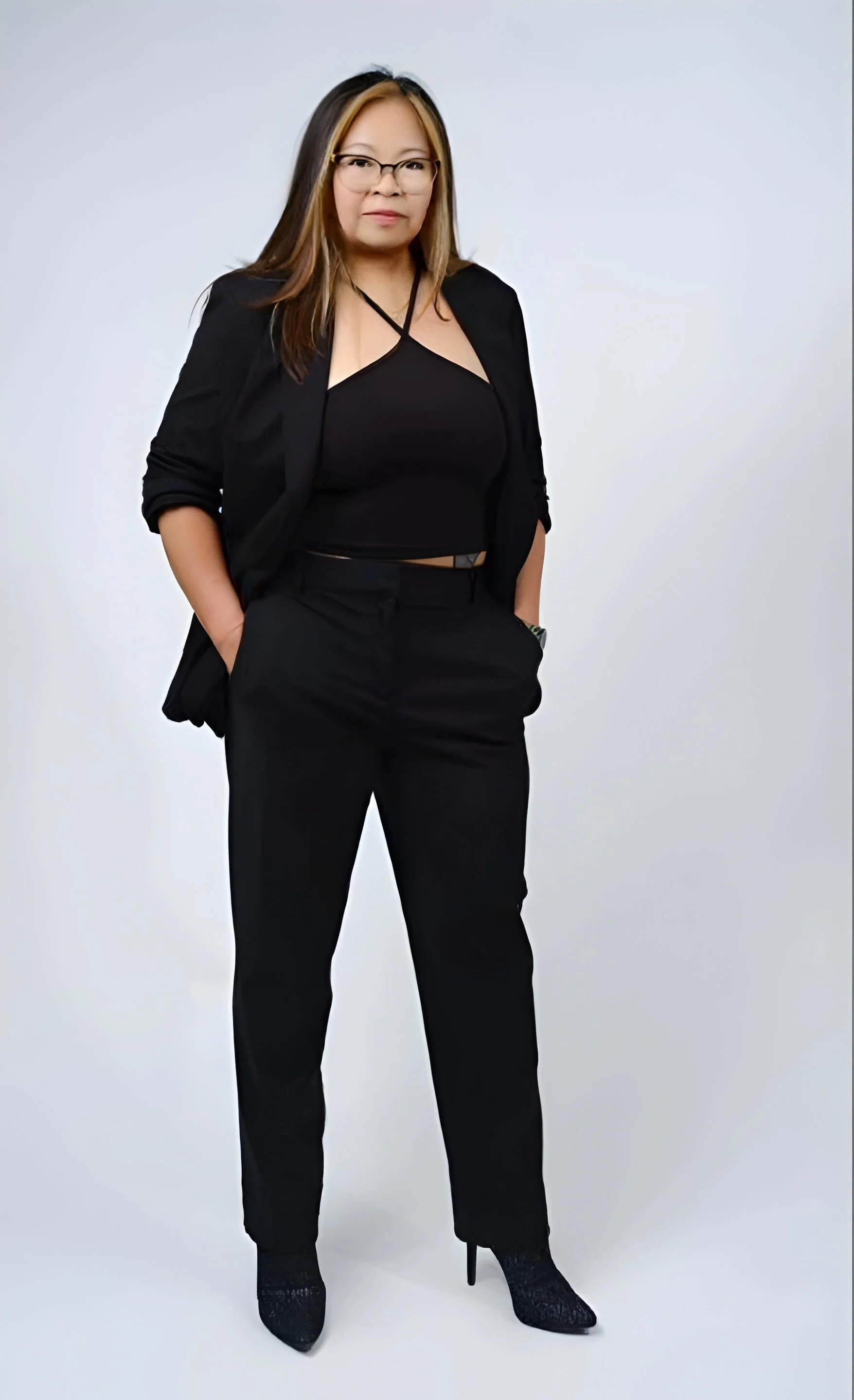 A woman in a black outfit, wearing glasses, standing with hands in her pockets, against a plain white background.