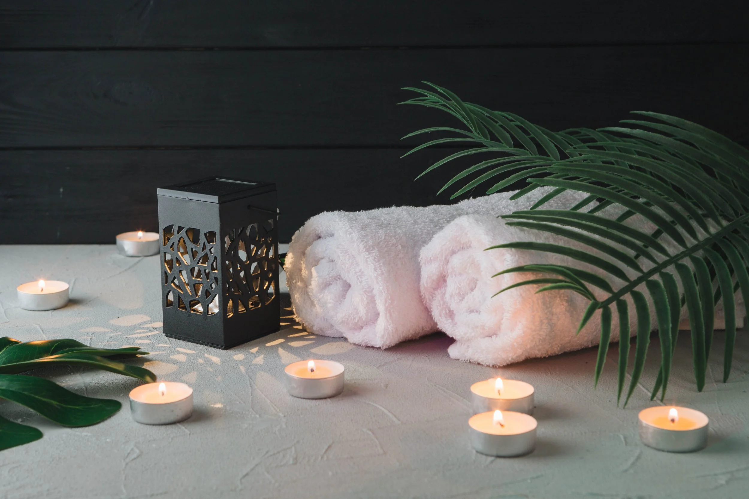Spa scene with rolled white towels, lit tea light candles, a decorative black lantern, and green tropical leaves against a dark background.