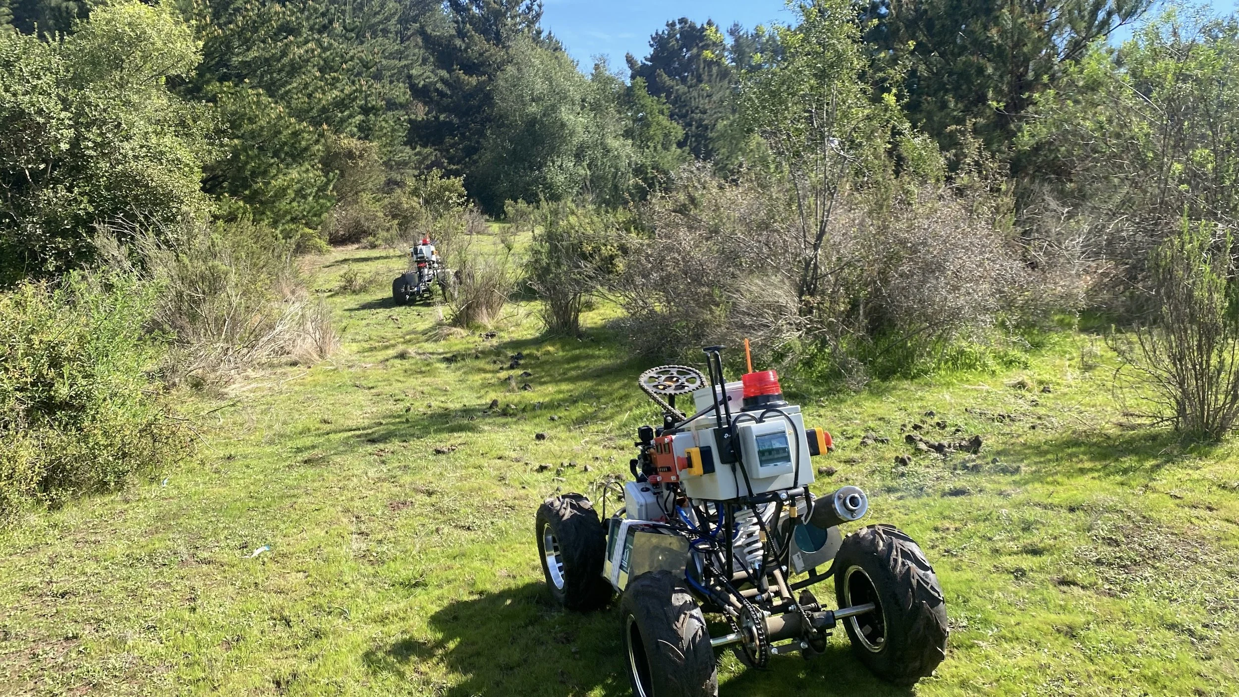 A small robotic vehicle with four large tires, mounted with a sensor and electronic components, is navigating a grassy trail in a wooded area. Another similar robot is visible further ahead on the trail.