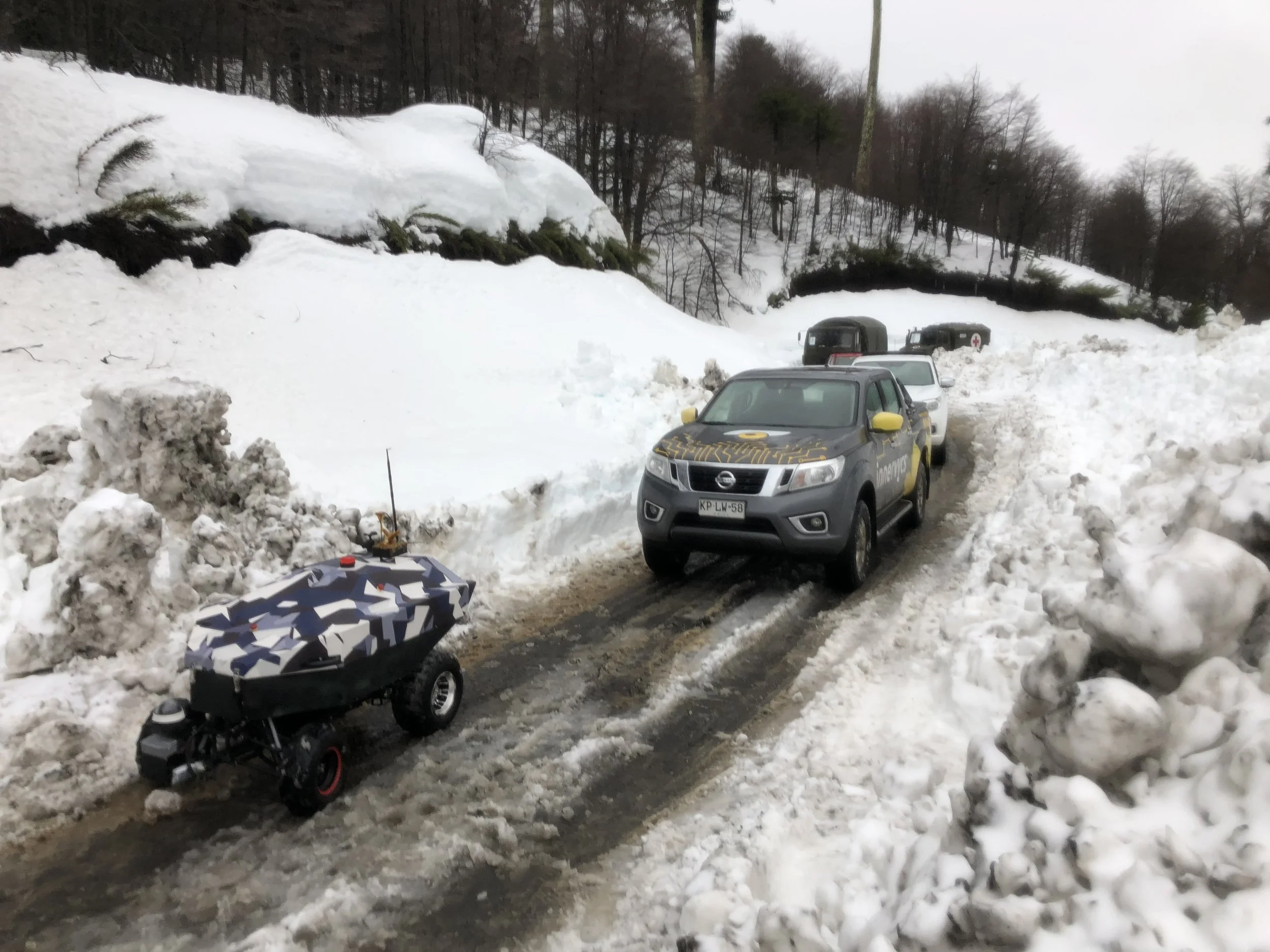 A snowy mountain road with police vehicles and a small autonomous robot vehicle on the street.