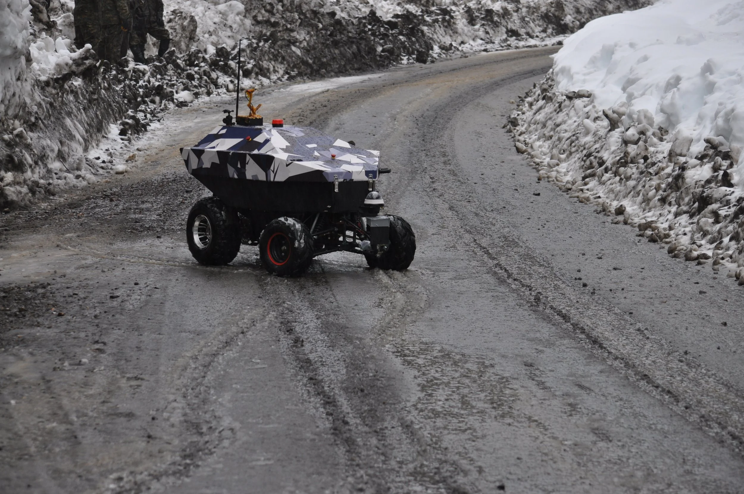 A small autonomous robotic vehicle driving on a wet, icy mountain road with snow on the sides.