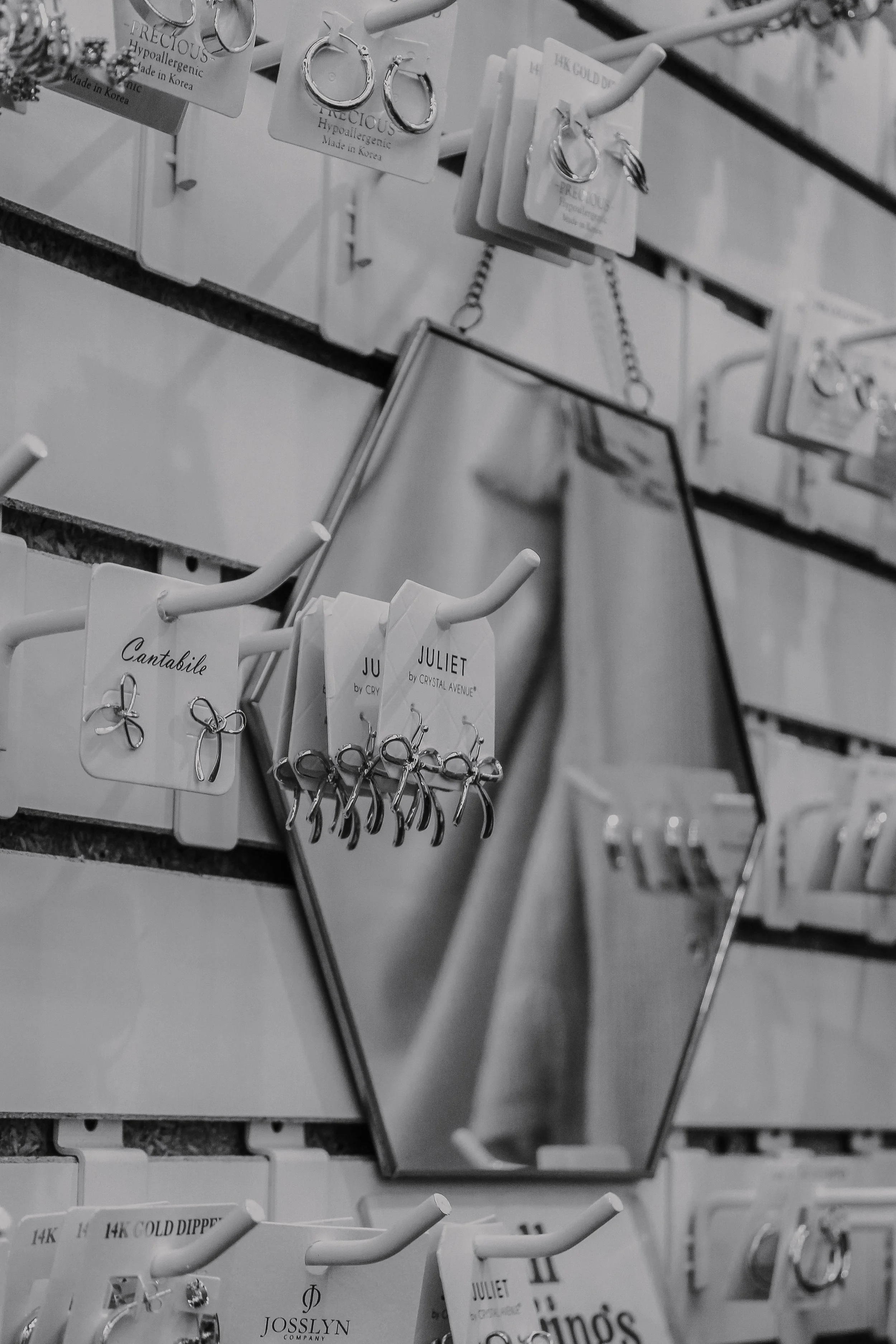 Display of assorted earrings on white cards, arranged on hooks and held by a mirror. The earrings vary in style, including hoops and bow designs. The display is part of a jewelry store, with some earrings labeled with brand names like Juliet and Cantabile.