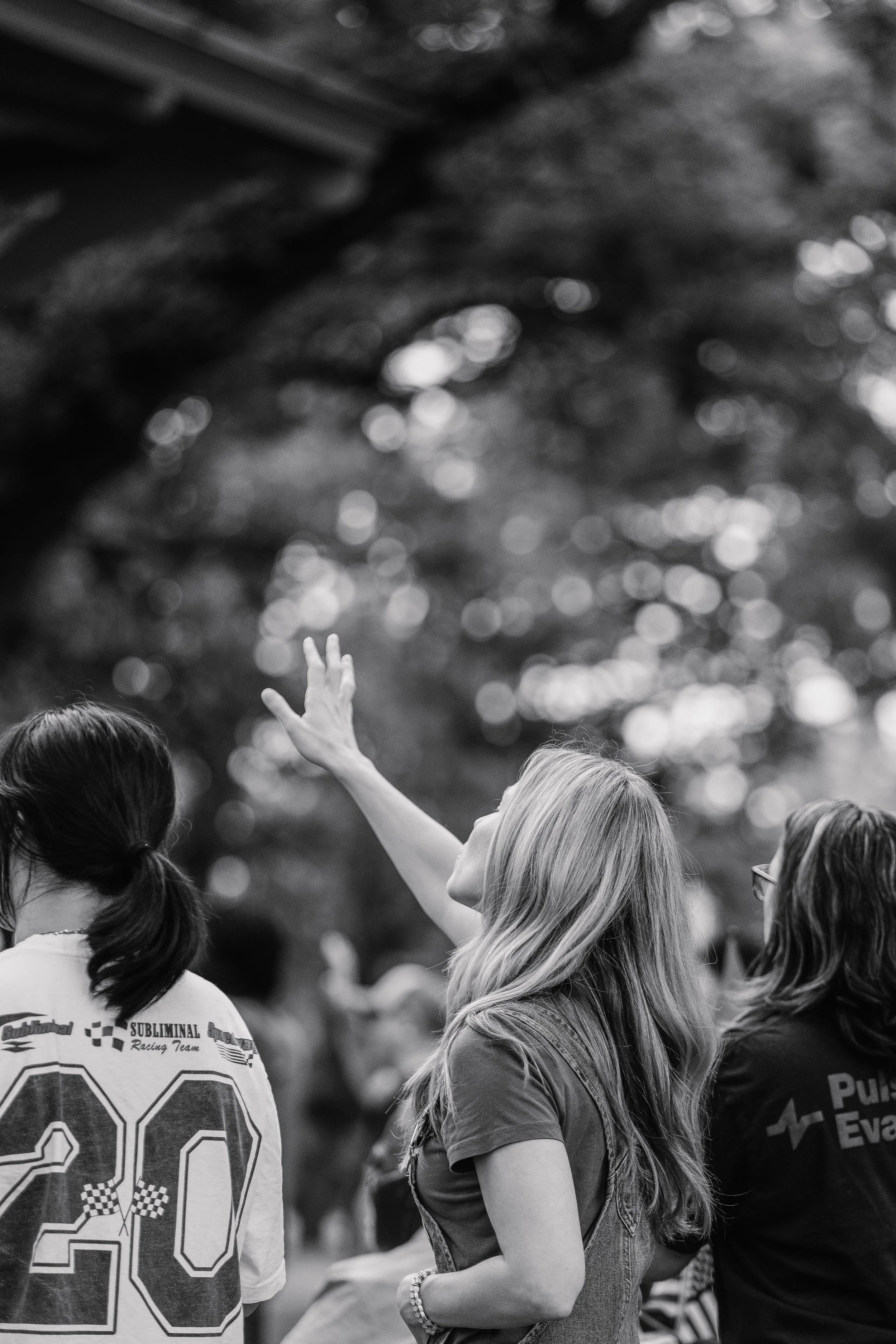 A woman with long blonde hair raising her hand, appears to be speaking or gesturing to a group of people outdoors. Two other women are visible, one with a ponytail wearing a sports jersey, and the other with glasses and curly hair. The background is blurred with trees, and the photo is in black and white.