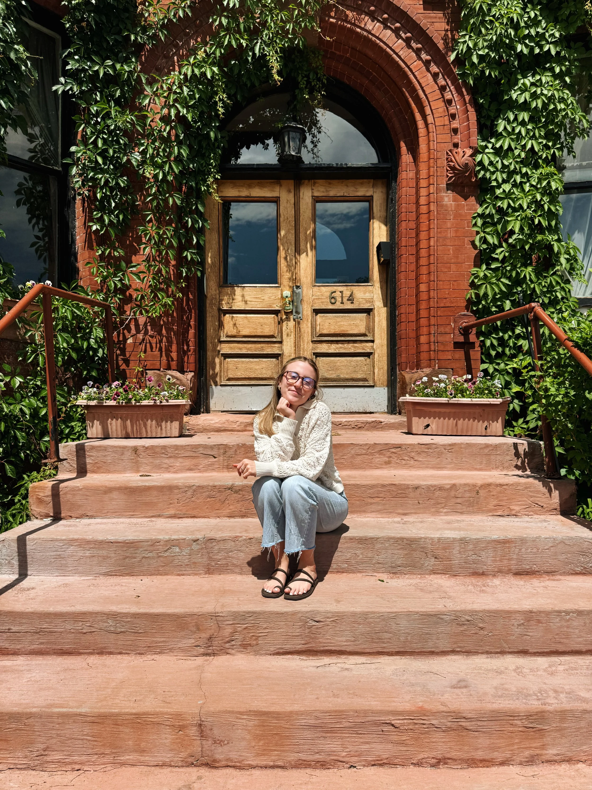 A woman sitting on the steps in front of a brick building with a wooden front door framed by green ivy and bushes. There are flower boxes on the steps, and the woman is smiling, wearing glasses, a white sweater, and light-wash jeans.