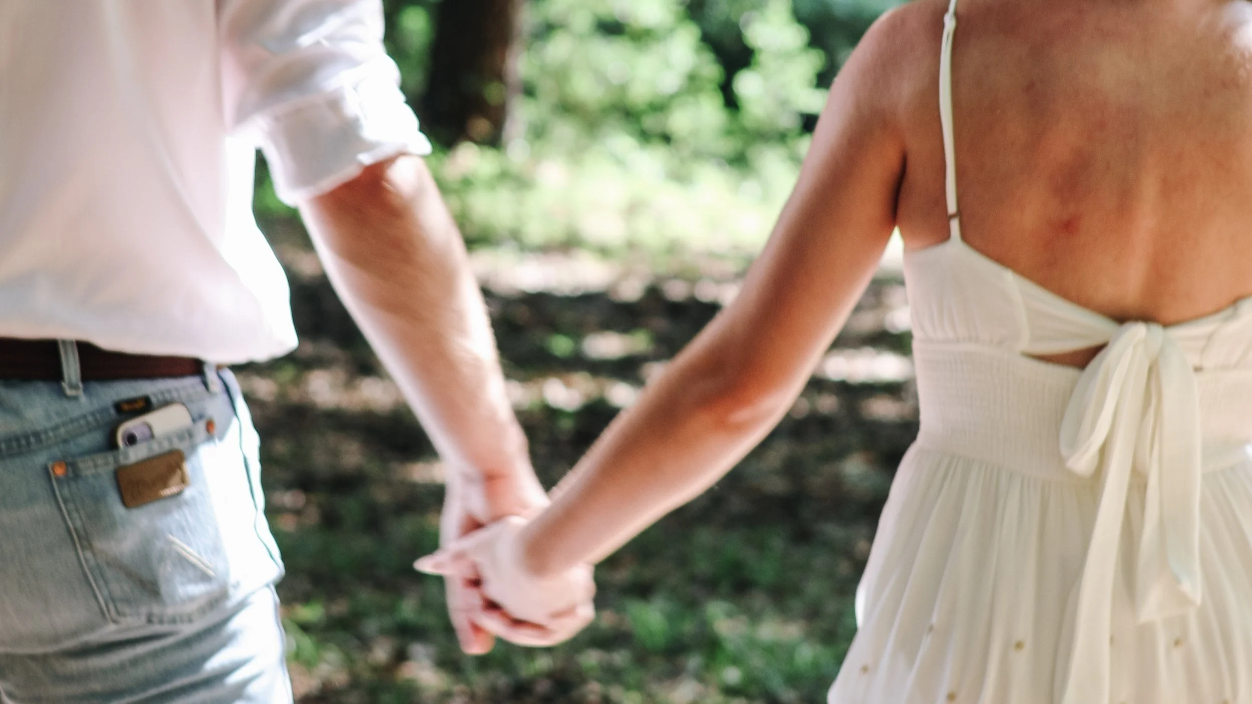 A couple holding hands outdoors in a wooded area, with only their torsos and hands visible.