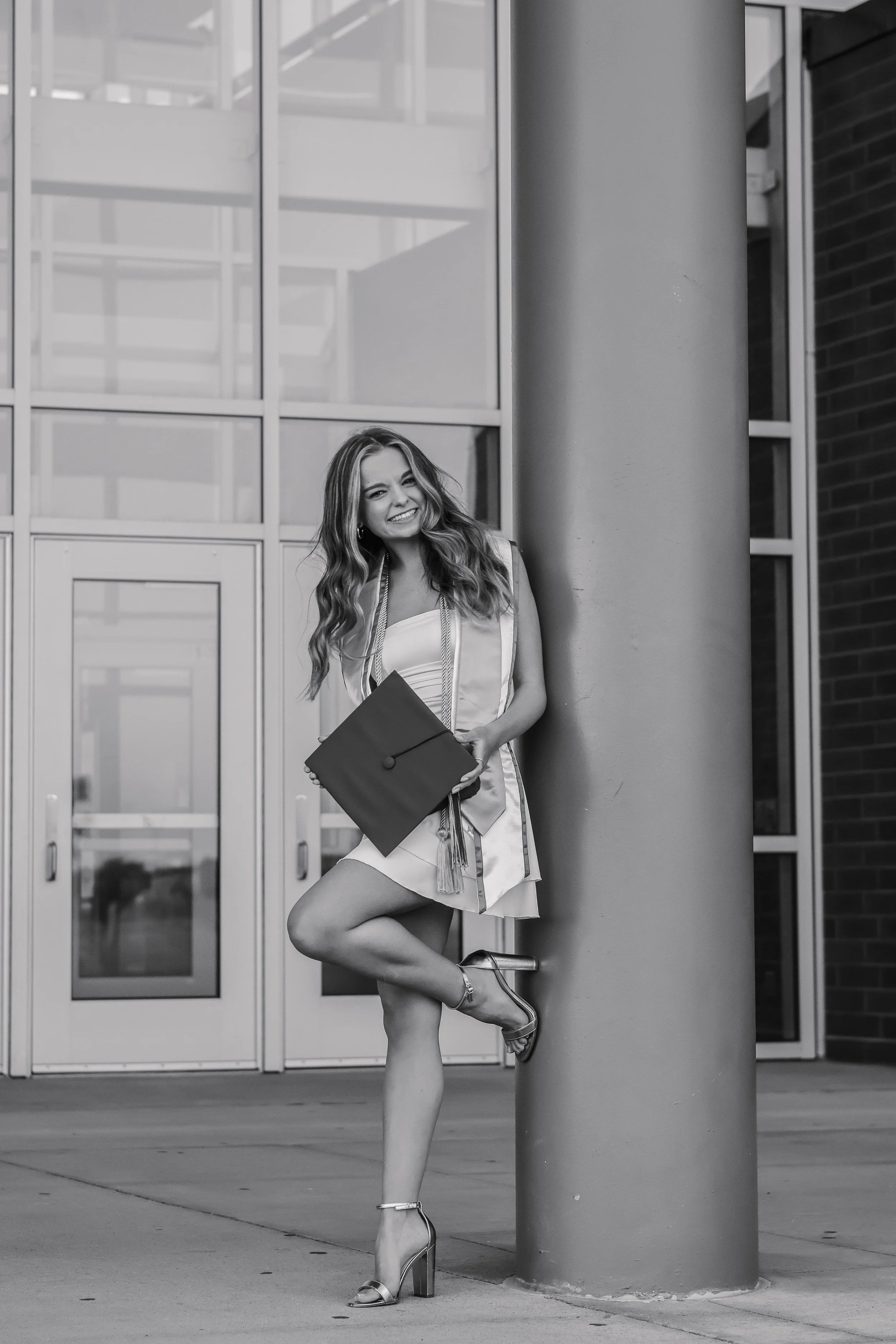 Black and white photo of a young woman in a graduation gown and cap, smiling and posing with one leg crossed in front of a modern building.