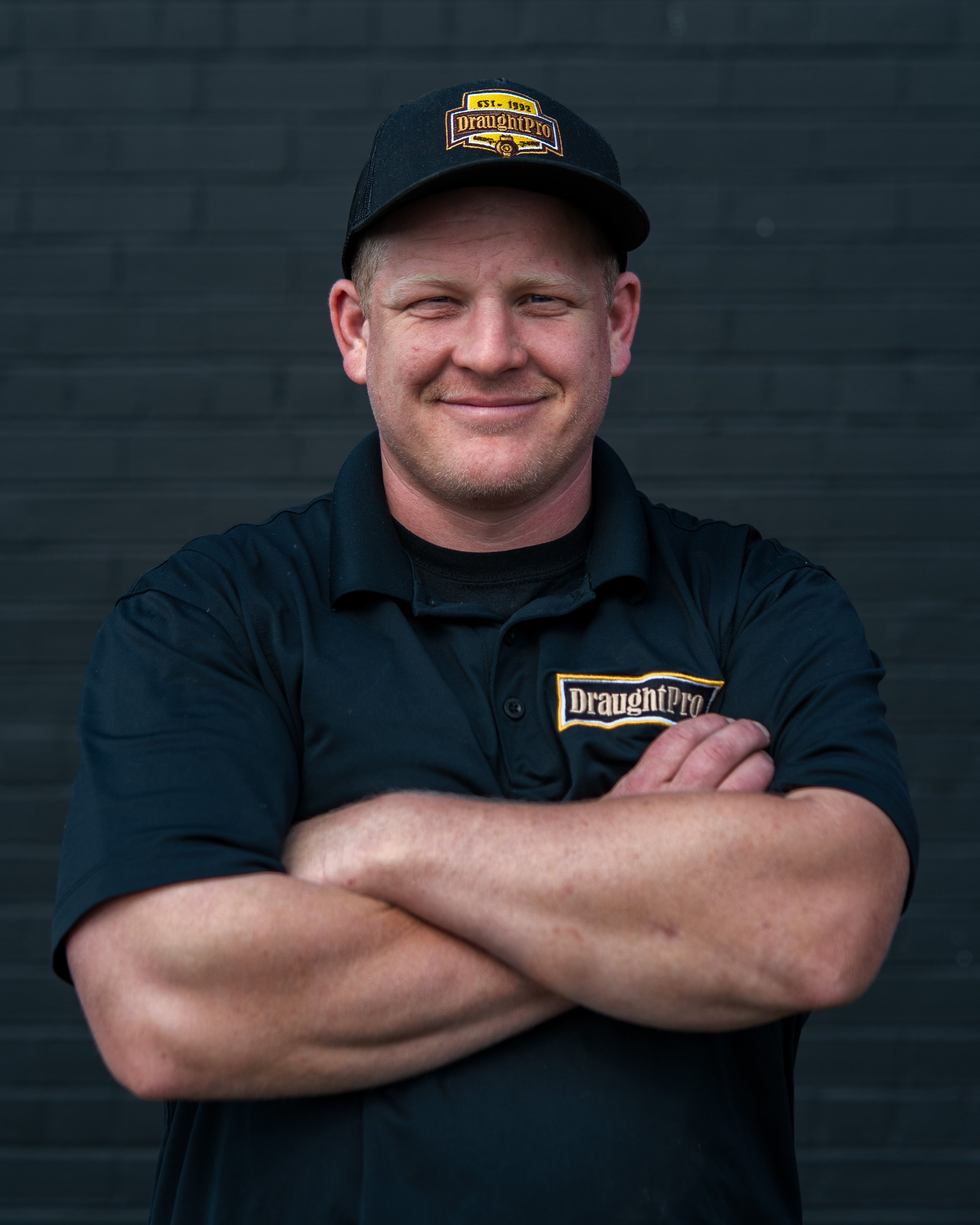 A man wearing a black cap and black shirt with a 'DraughtPro' logo, standing in front of a dark wall, smiling with his arms crossed.