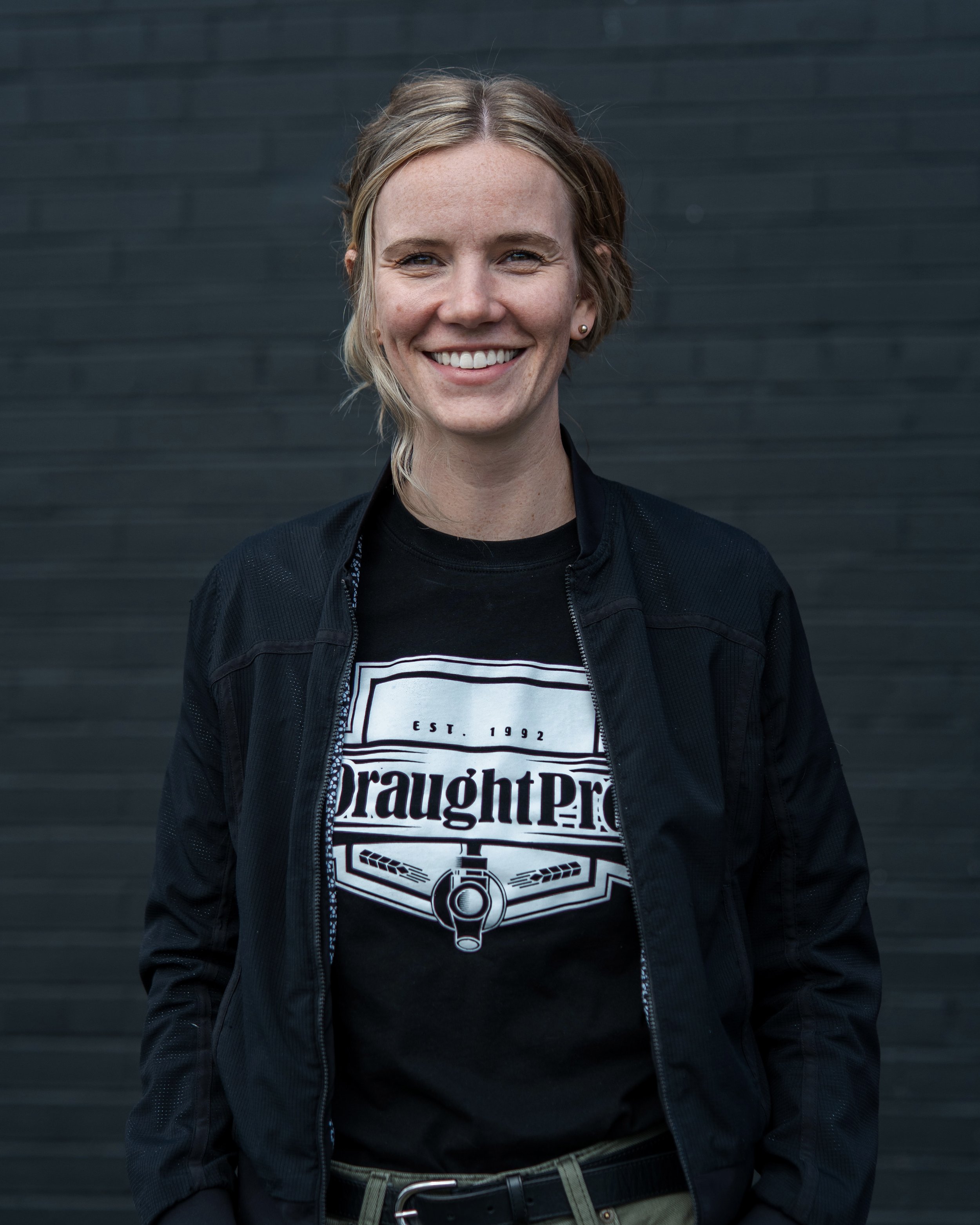 A smiling woman with short blonde hair, wearing a black jacket over a black T-shirt with a graphic design, standing in front of a dark gray brick wall.