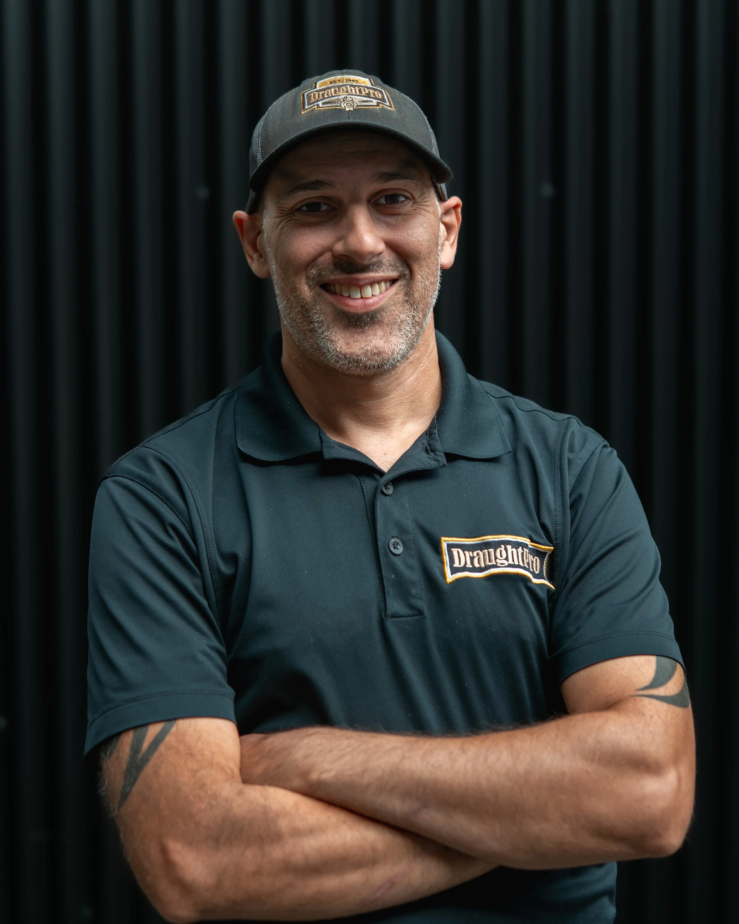 A man smiling with crossed arms, wearing a black Drewnoff polo shirt and cap, standing in front of a black curtain background.